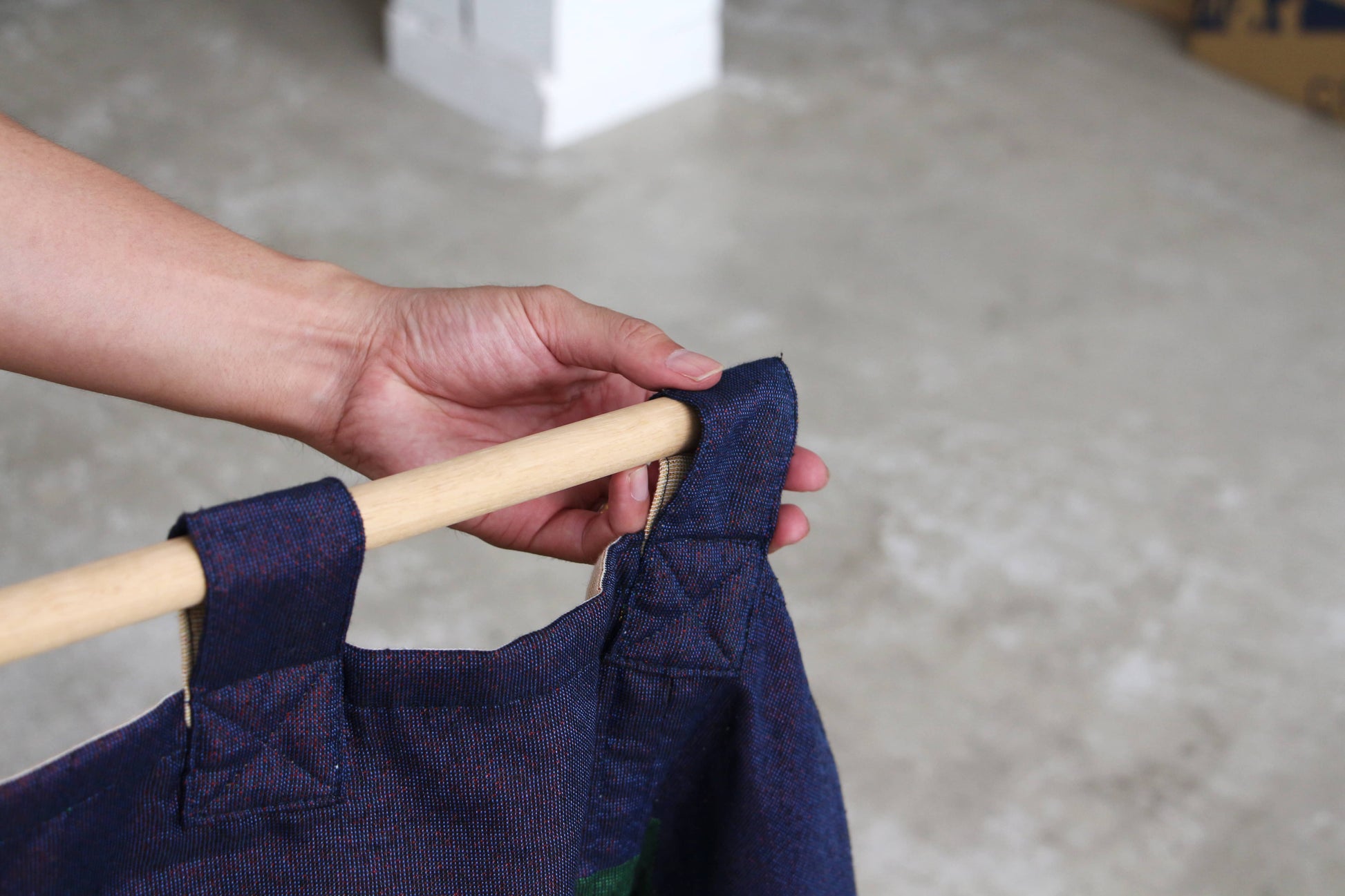 A hand holds the DETAIL Noren Curtain by DETAIL, with a wooden rod threaded through fabric loops on a blue garment featuring subtle kimono patterns, set against a concrete floor background.