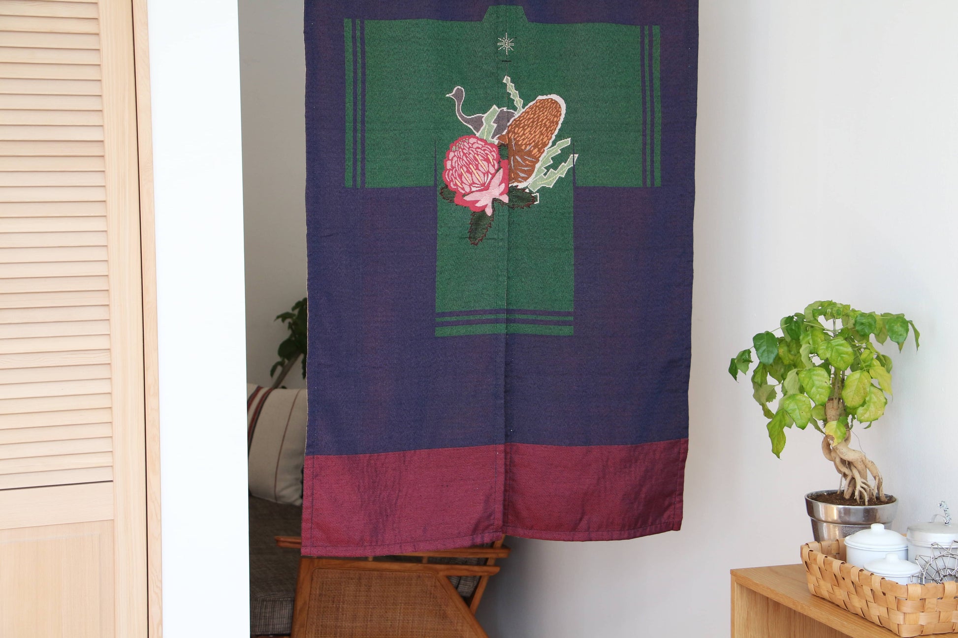 The DETAIL Noren Curtain by DETAIL, featuring kimono patterns and floral motifs, hangs in the doorway. A small table with a potted plant and teaware sits against a white wall in this cozy, well-lit room.