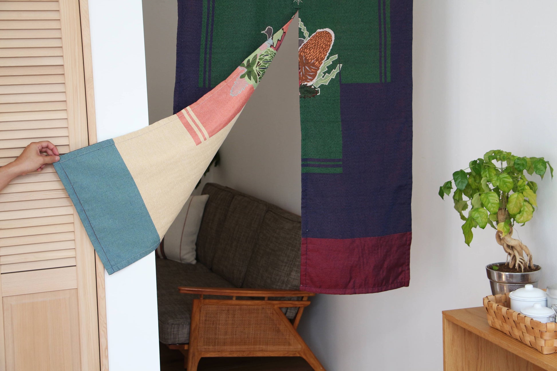 A hand pulls back the DETAIL Noren Curtain by DETAIL, revealing a cozy living room with a wooden chair, gray cushion, and a green leafy plant atop a small wooden cabinet.