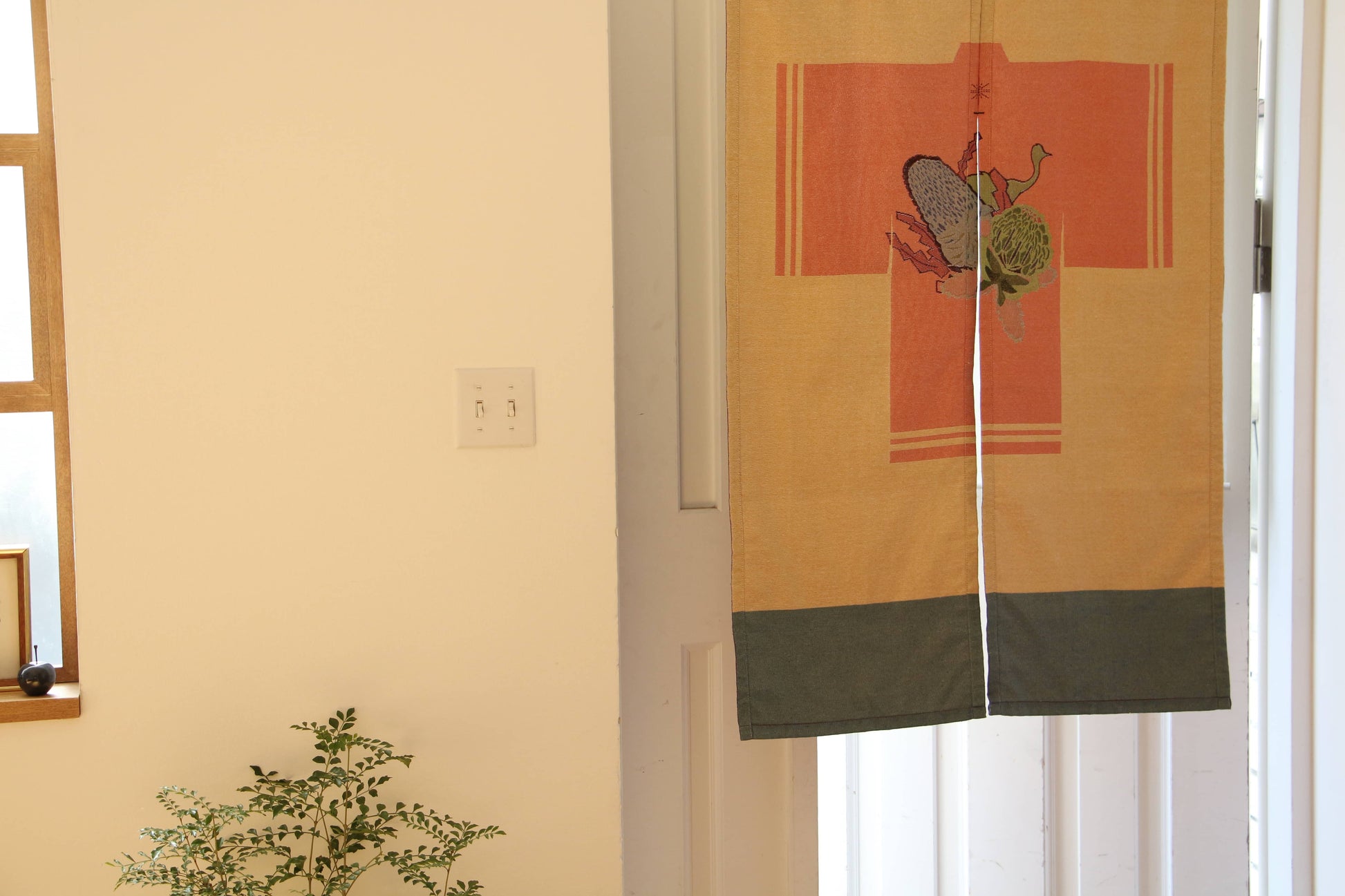 A brightly lit room features a potted plant near light switches and a beige DETAIL Noren Curtain by DETAIL, decorated with kimono patterns and insect motifs, hanging in the doorway.