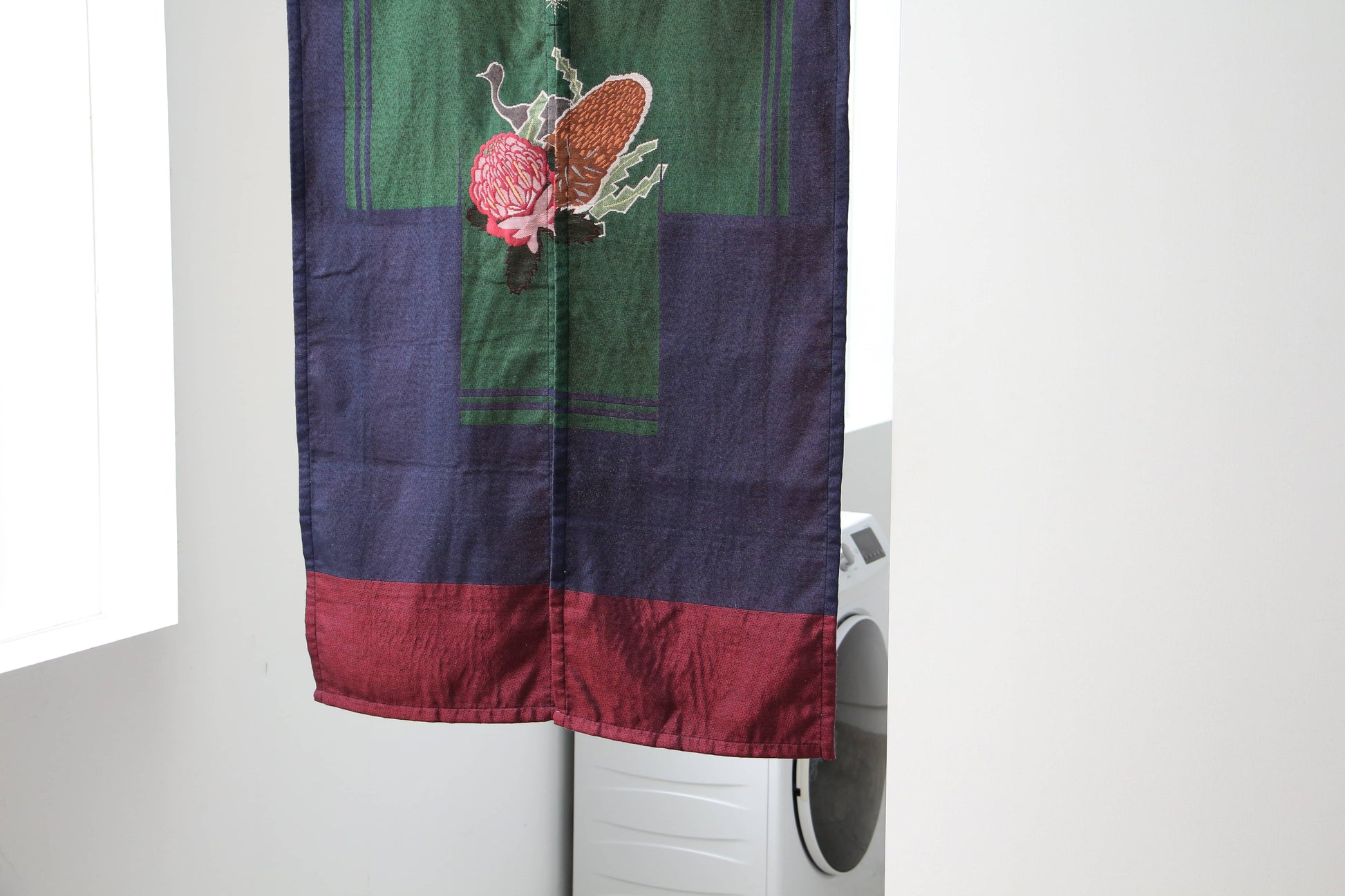 The DETAIL Noren Curtain by DETAIL, in dark blue and maroon with a mushroom and fern pattern, partially covers a white washing machine in a bright, minimalist room for a subtle, plant-inspired accent.