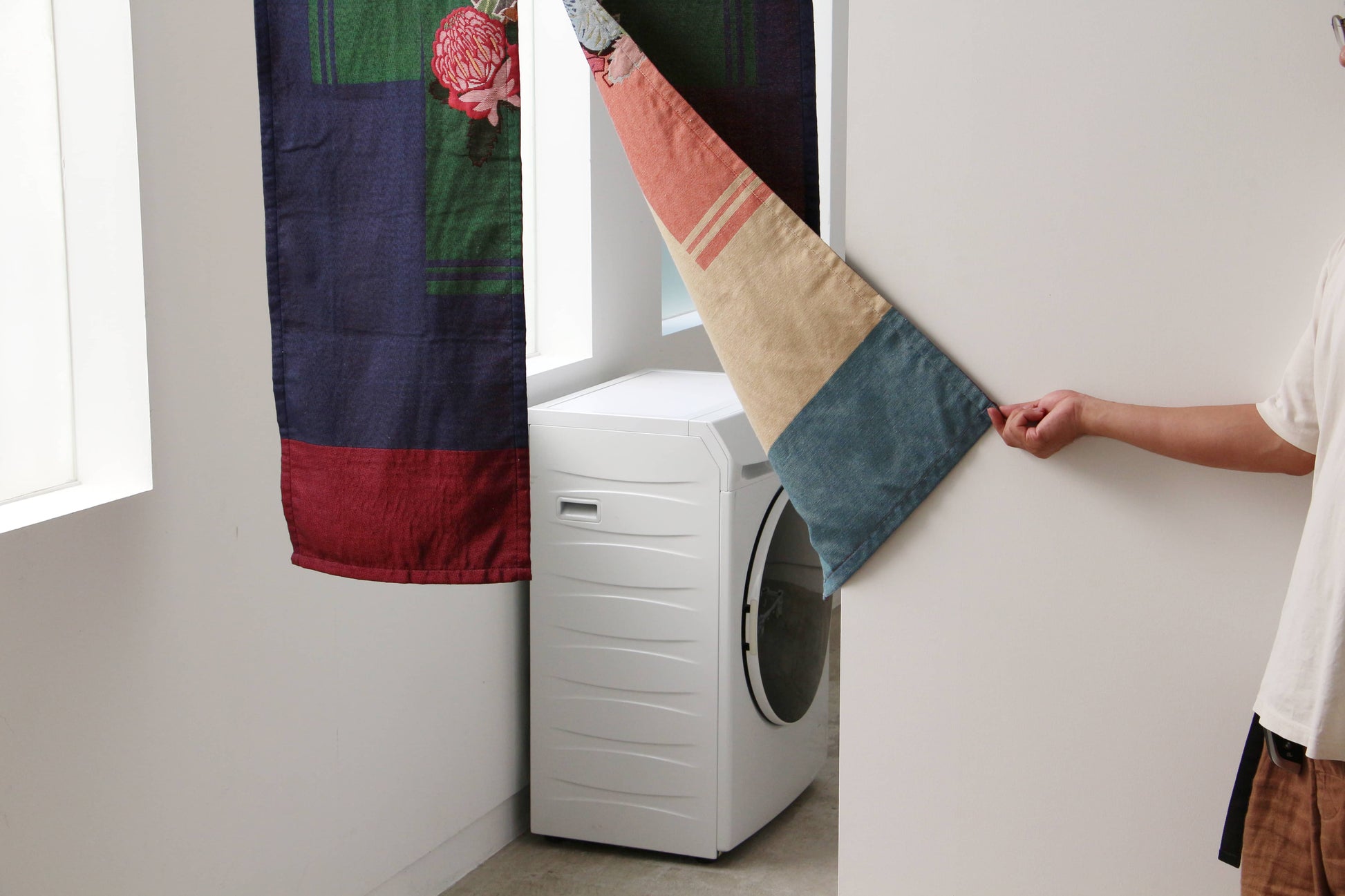A person pulls back the DETAIL Noren Curtain by DETAIL, featuring kimono patterns, to reveal a white front-loading washing machine in a bright, minimalist room.