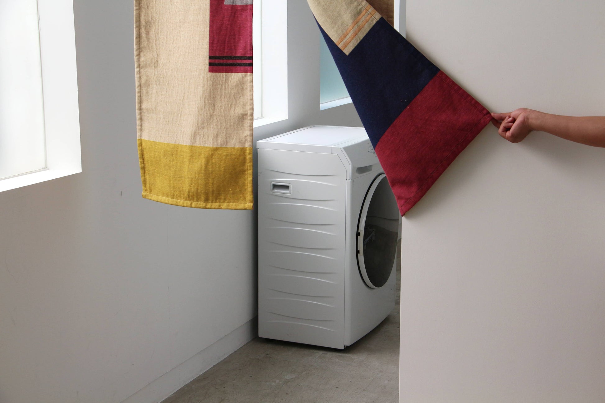 A white front-loading washing machine stands near two windows, partially covered by a DETAIL Noren Curtain from DETAIL with kimono patterns as a hand pulls back one fabric panel.
