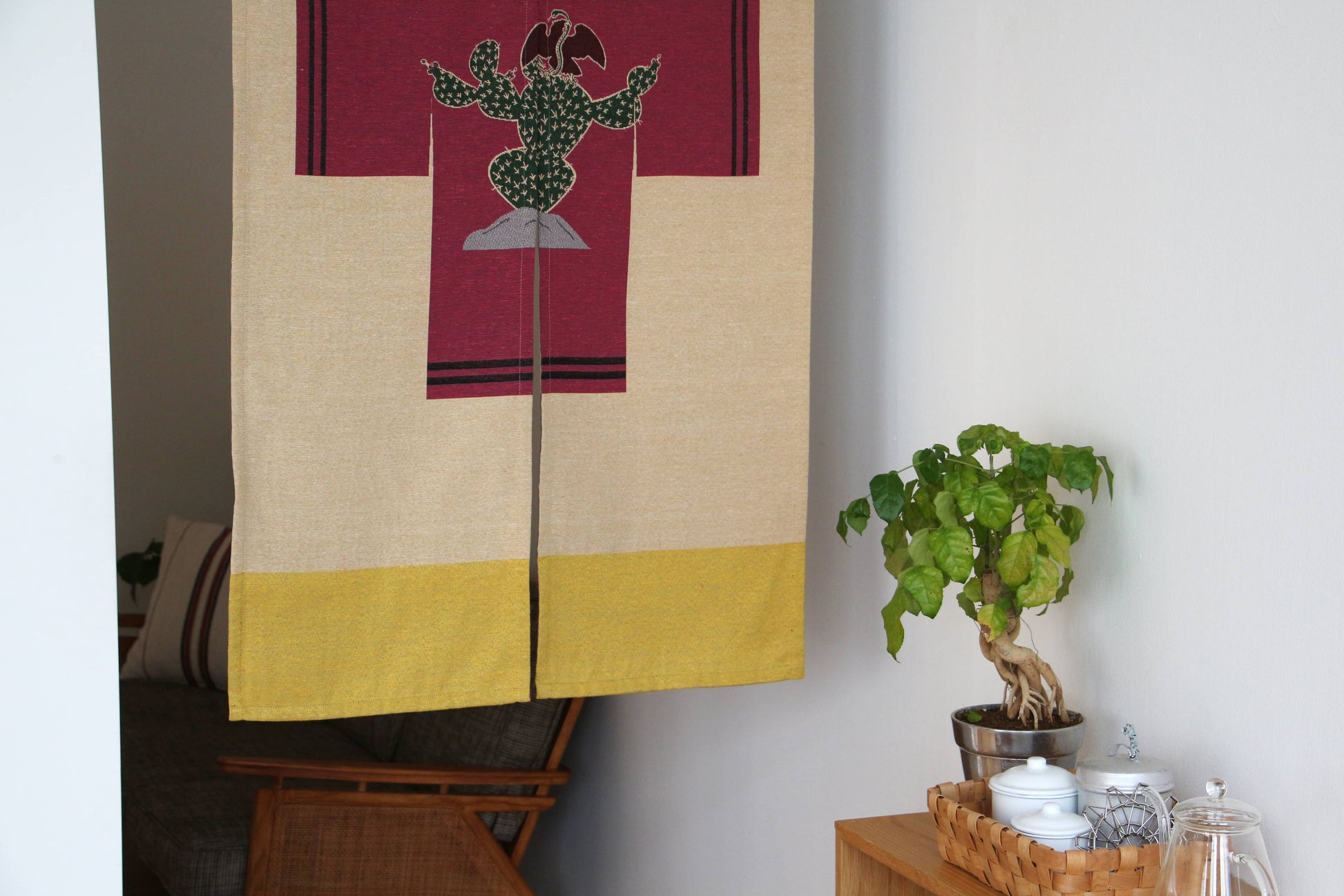 A cozy room features a wooden chair, cushion, and table. In the foreground hangs the DETAIL Noren Curtain by DETAIL with a cactus design. A side table holds a small plant and a basket with jars and a teapot.