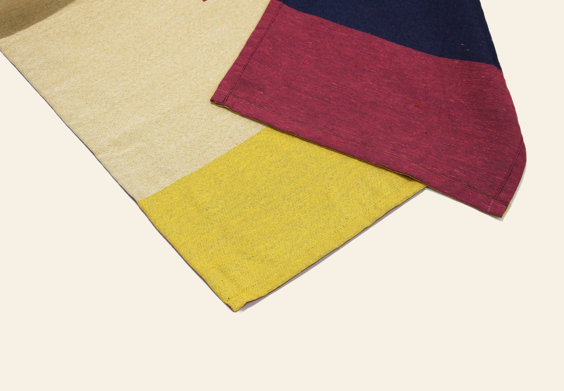 Two DETAIL Noren Curtain fabrics overlap on a light background—one with yellow and beige panels, the other with dark blue, red, and pink sections plus visible stitching. Their design recalls kimono patterns and traditional Noren curtains by DETAIL.