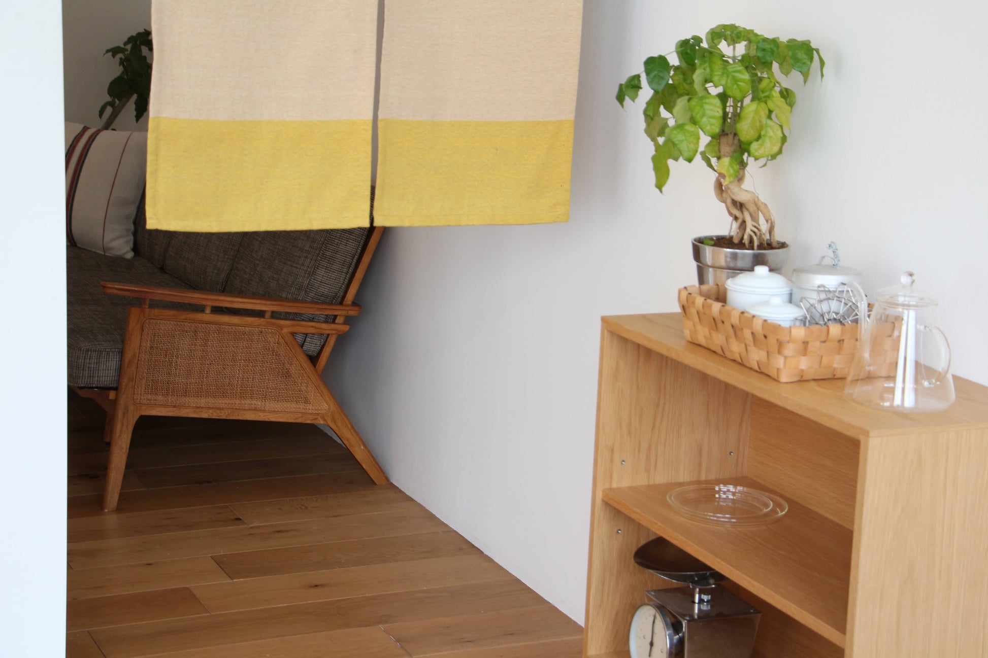 A wooden armchair sits in a cozy corner, partially hidden by the DETAIL Noren Curtain from DETAIL. A shelf holds a potted plant, teapot, and kitchenware. Wooden floors and simple decor complete the warm, welcoming vibe.