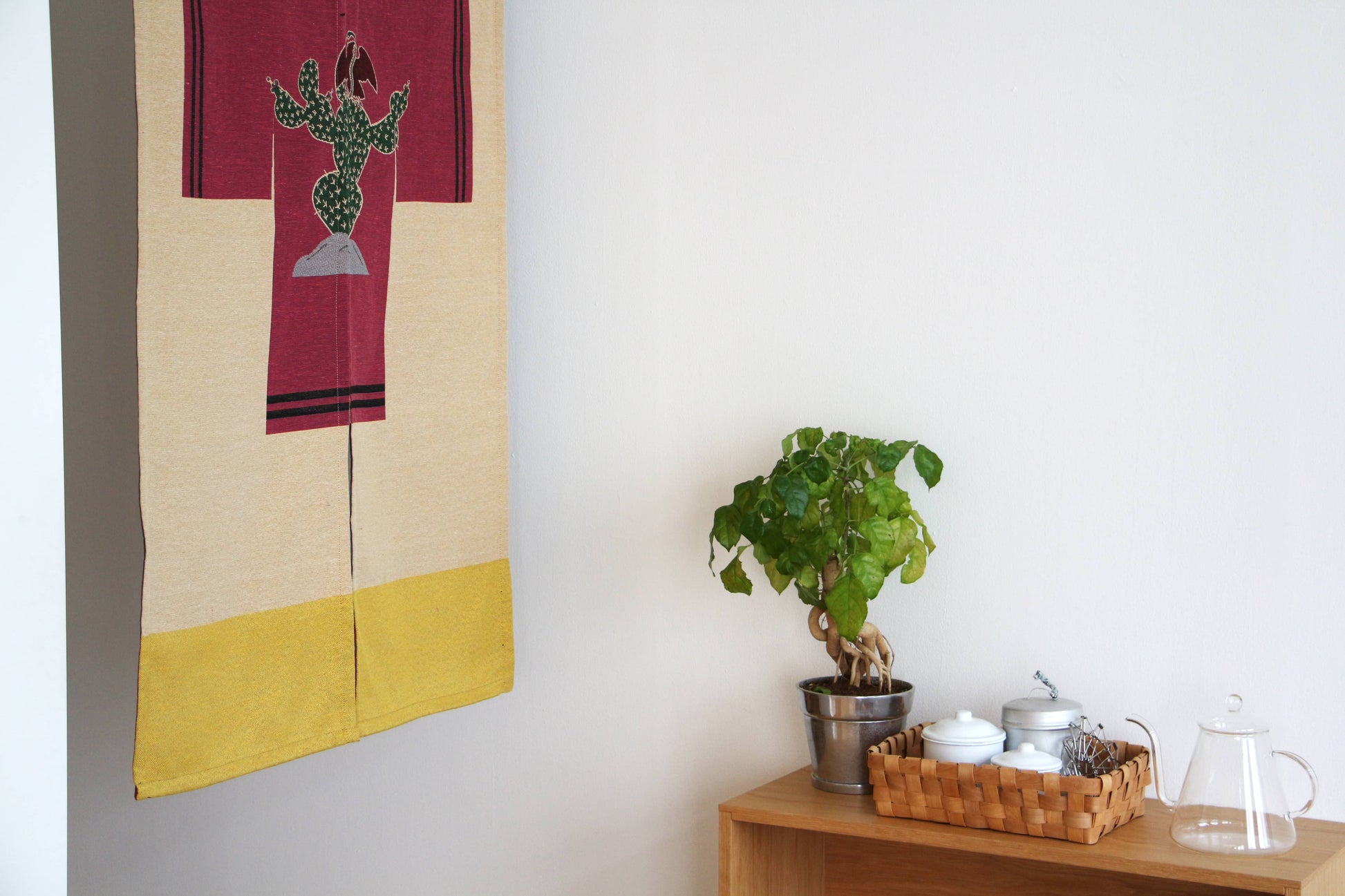 A small indoor plant in a silver pot sits on a shelf beside a basket of teapots and cups, with a glass teapot nearby. Above, a DETAIL Noren Curtain by DETAIL featuring a colorful cactus design decorates the white wall.