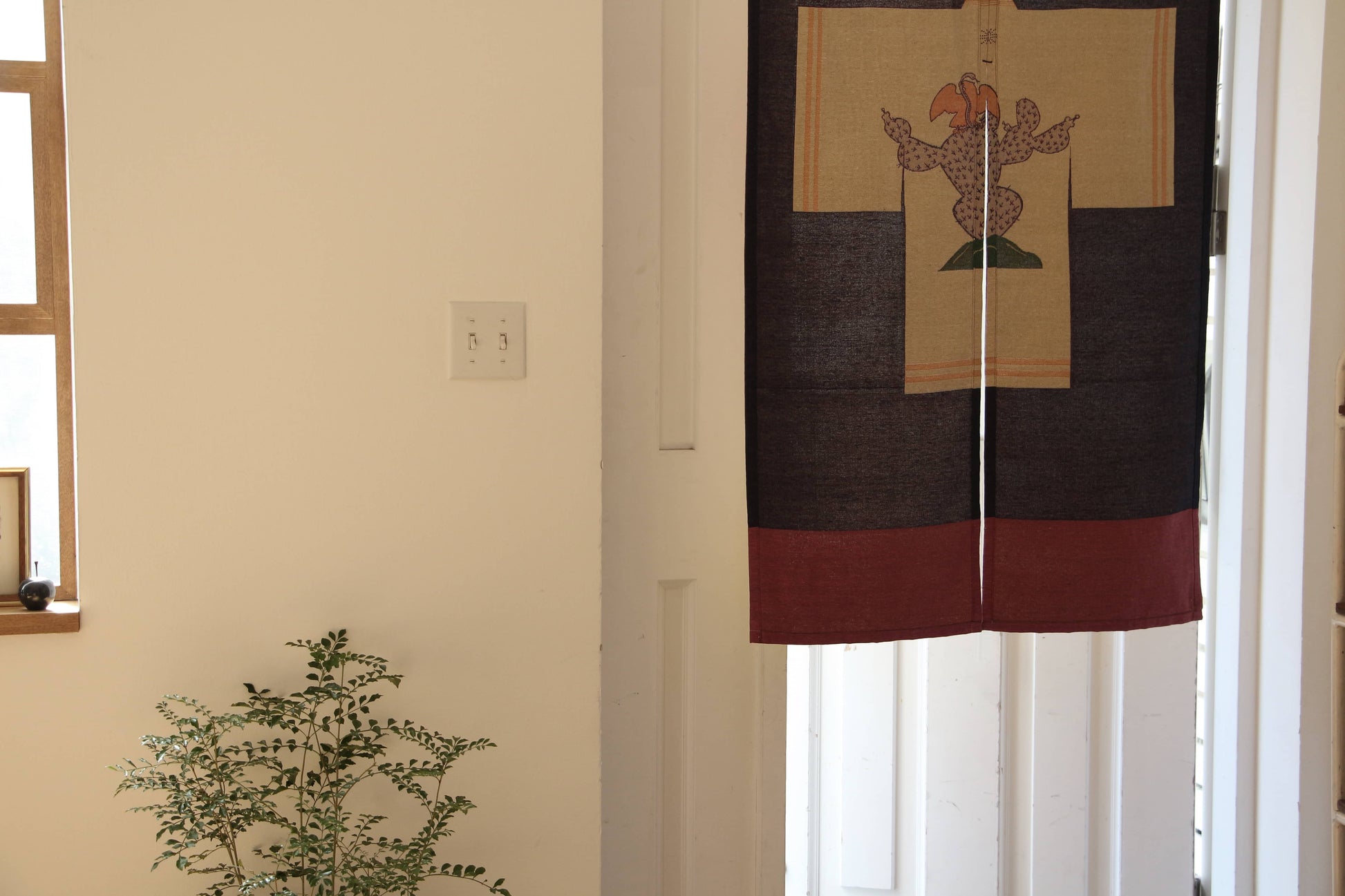 A doorway features the DETAIL Noren Curtain by DETAIL, showcasing a vibrant animal design inspired by kimono patterns. Nearby, a small green potted plant sits as sunlight filters through the window on the left.