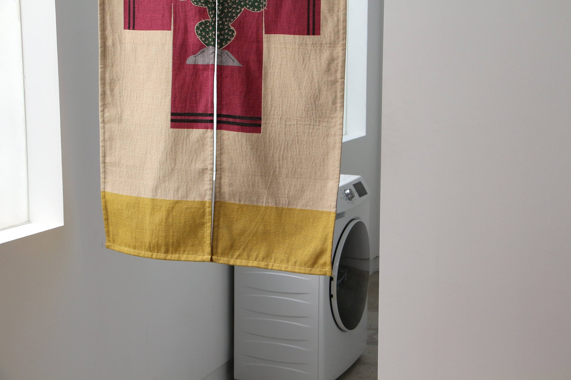 A DETAIL Noren Curtain by DETAIL, featuring a cheerful cactus design in yellow, beige, and red sections, partially conceals a washing machine in a bright, minimalistic room.