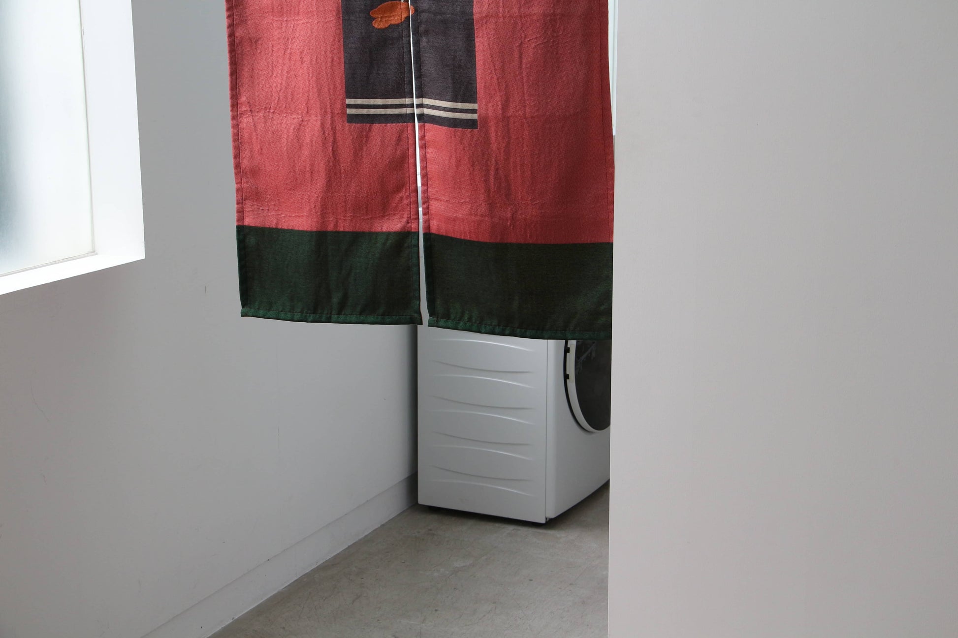 A DETAIL Noren Curtain by DETAIL, adorned with kimono patterns, hangs in a minimalistic white room with a concrete floor; a washing machine is partially visible behind it.
