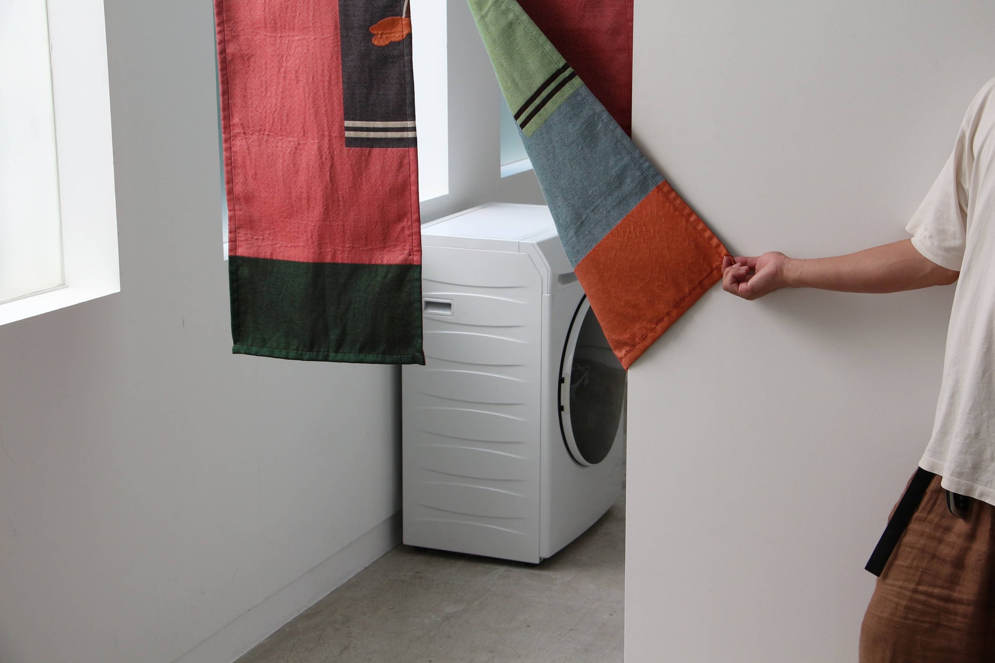A person in a white shirt pulls aside the DETAIL Noren Curtain by DETAIL, featuring vivid kimono patterns, to reveal a white front-loading washing machine in a bright, minimal laundry room.