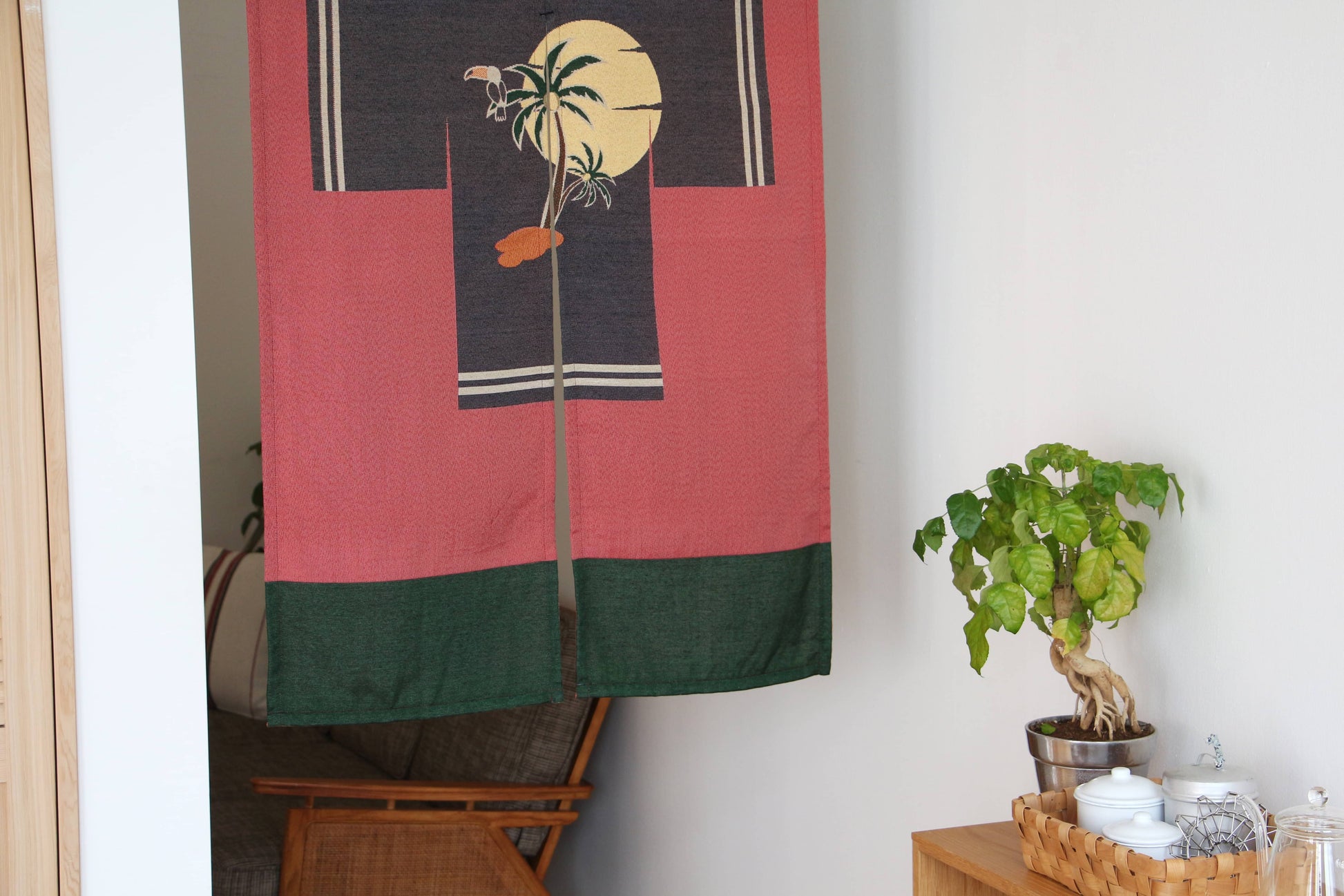 The DETAIL Noren Curtain by DETAIL, with a tropical palm tree and sun design, hangs in the doorway. Nearby, a small table holds a potted plant, wicker basket with teapot and cups, and a wooden chair is partially visible.