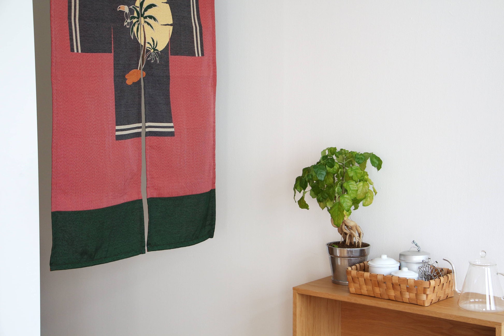 A minimalist room corner with a wooden shelf holding a potted plant, a wicker tray with white containers, and the DETAIL Noren Curtain by DETAIL featuring a sun and palm tree design hanging on the wall.