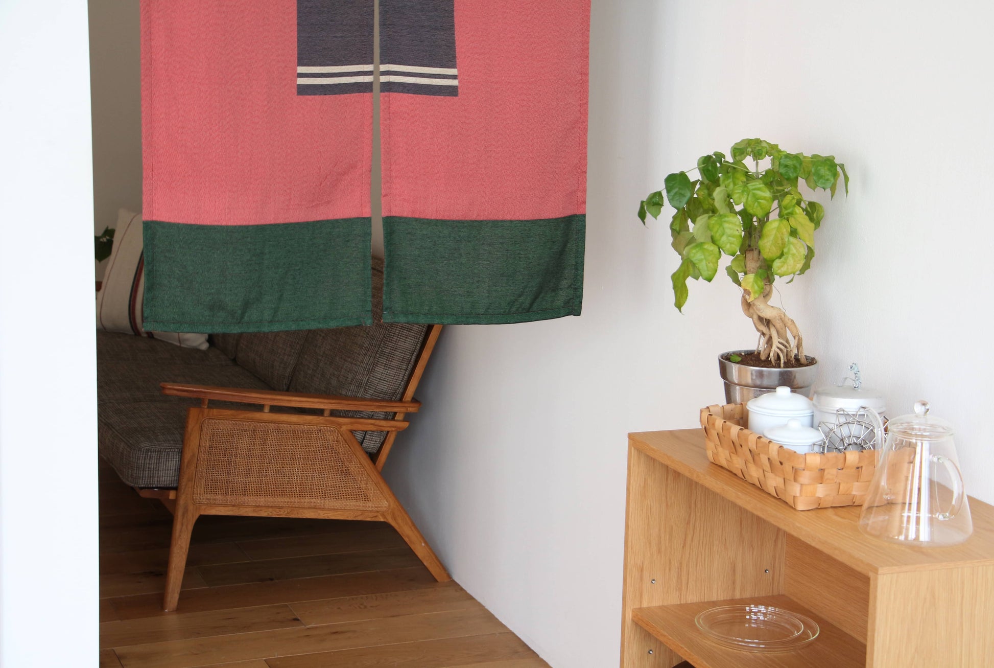 A cozy corner with a wooden armchair, partially hidden by the DETAIL Noren Curtain in pink and green, a potted plant on a shelf, and a woven basket with teapots and cups on a light wooden cabinet.