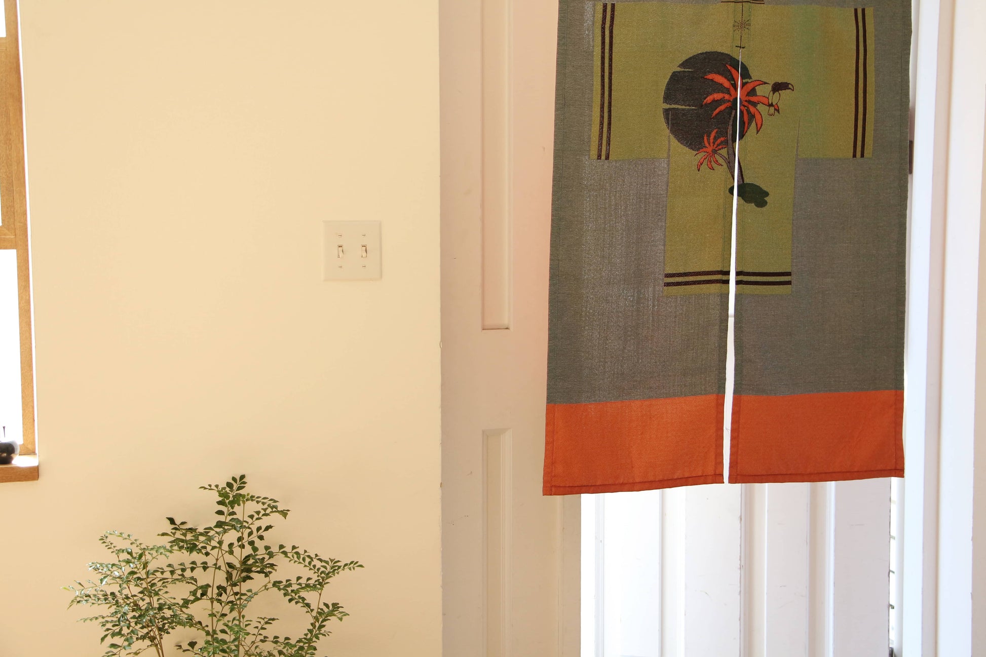 A small leafy potted plant sits on the floor by a white wall and a wooden-framed window. A doorway is partially covered by the DETAIL Noren Curtain from DETAIL, featuring orange, green, and black kimono patterns.