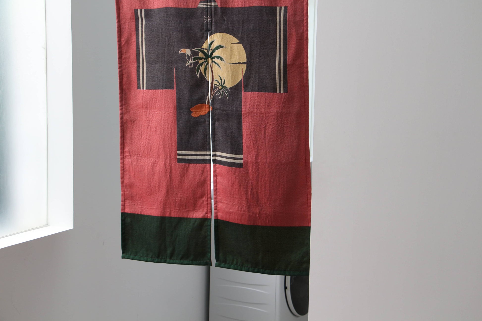 The DETAIL Noren Curtain by DETAIL features red and black panels with a yellow moon, palm trees, and a bird silhouette, its plant-inspired design softly concealing a white washing machine in the doorway.