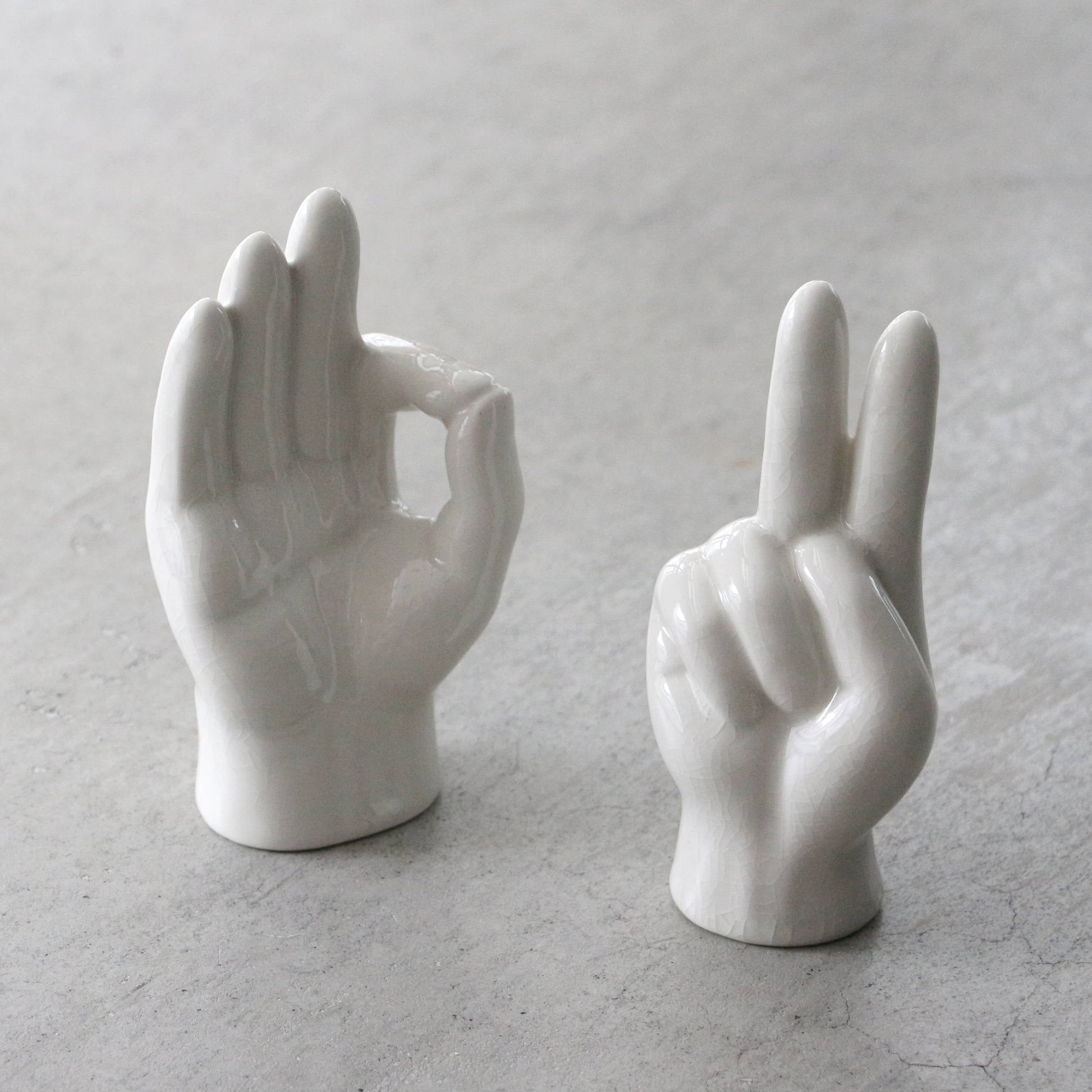 The DETAIL Porcelain Hand Peace by DETAIL features two white ceramic hands—one with an OK gesture, the other a peace sign—resting on a light gray surface, ideal as unique accessory stands or striking decor.
