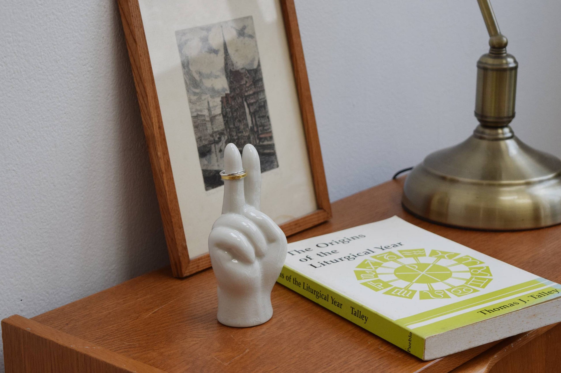 A DETAIL Porcelain Hand Peace accessory stand by DETAIL holding two rings rests on a wooden table beside The Origins of the Liturgical Year, a brass lamp, and framed black-and-white artwork.