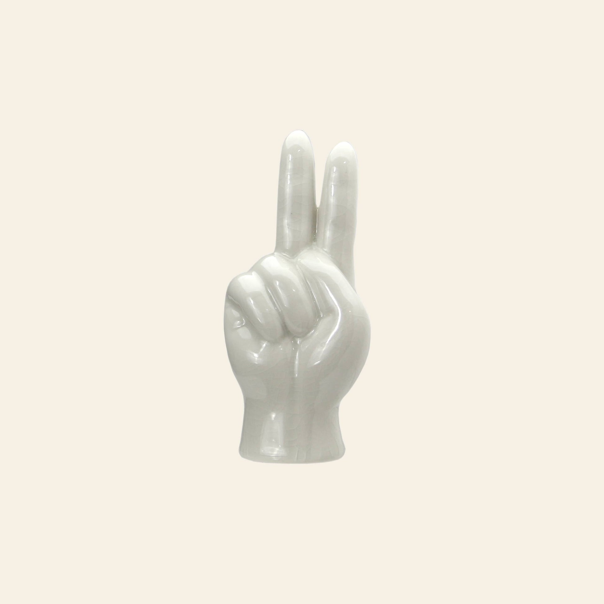 The DETAIL Porcelain Hand Peace sculpture by DETAIL features a glossy white finish and raised fingers, set against a beige background—an eye-catching ceramic accent ideal as an accessory stand or unique decorative piece.