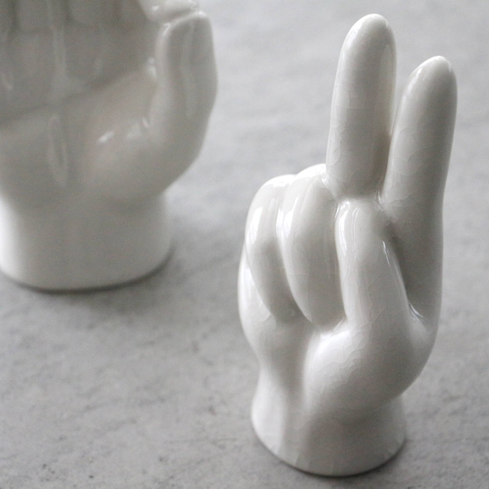 The DETAIL Porcelain Hand Peace by DETAIL displays a victory sign and doubles as a chic accessory stand. Another similar ceramic piece appears partially in the background on a gray surface.