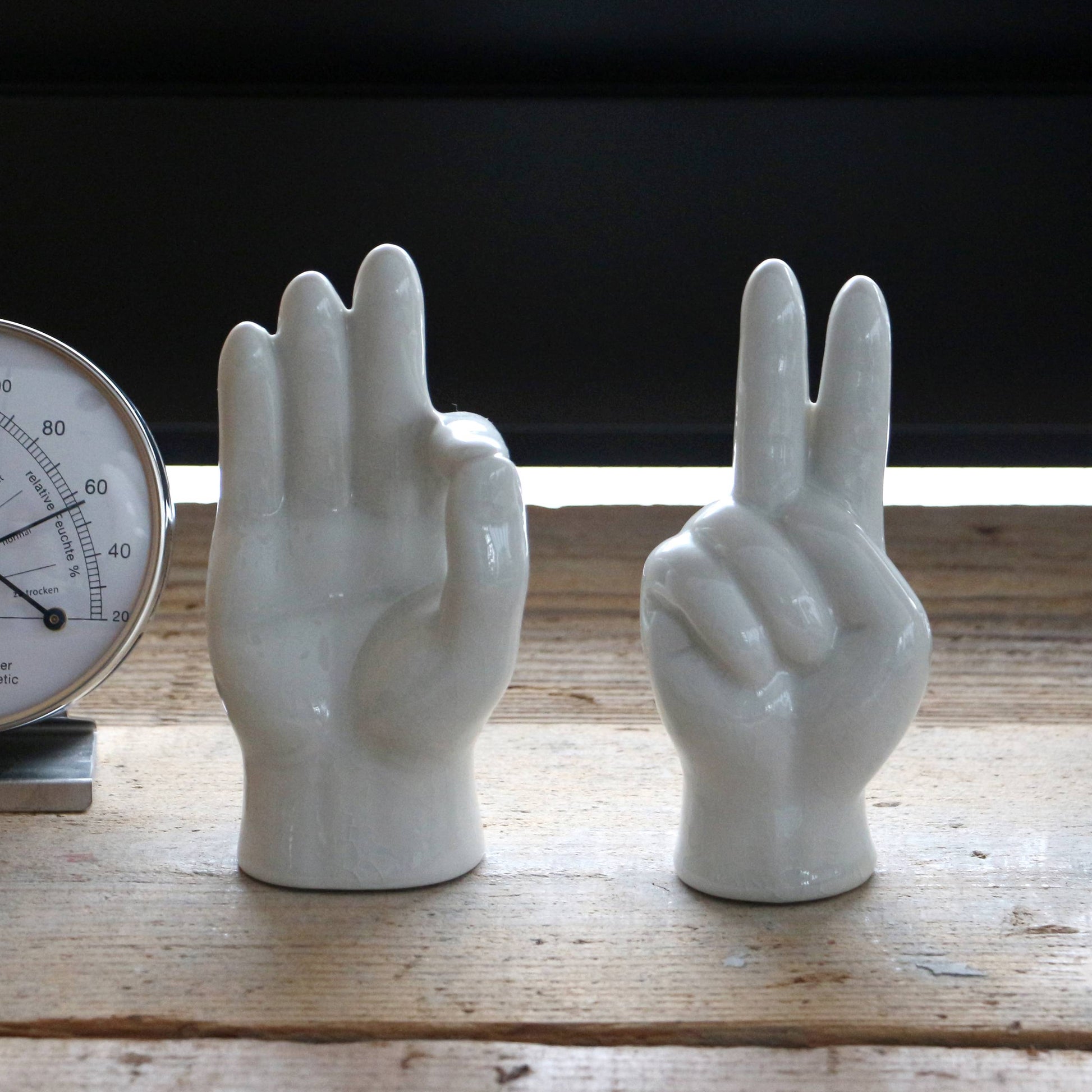 Two white ceramic hand sculptures by DETAIL rest on a wooden surface—one forms the OK gesture, the other displays the Porcelain Hand Peace sign from DETAIL. A partially visible thermometer and accessory stand appear on the left.