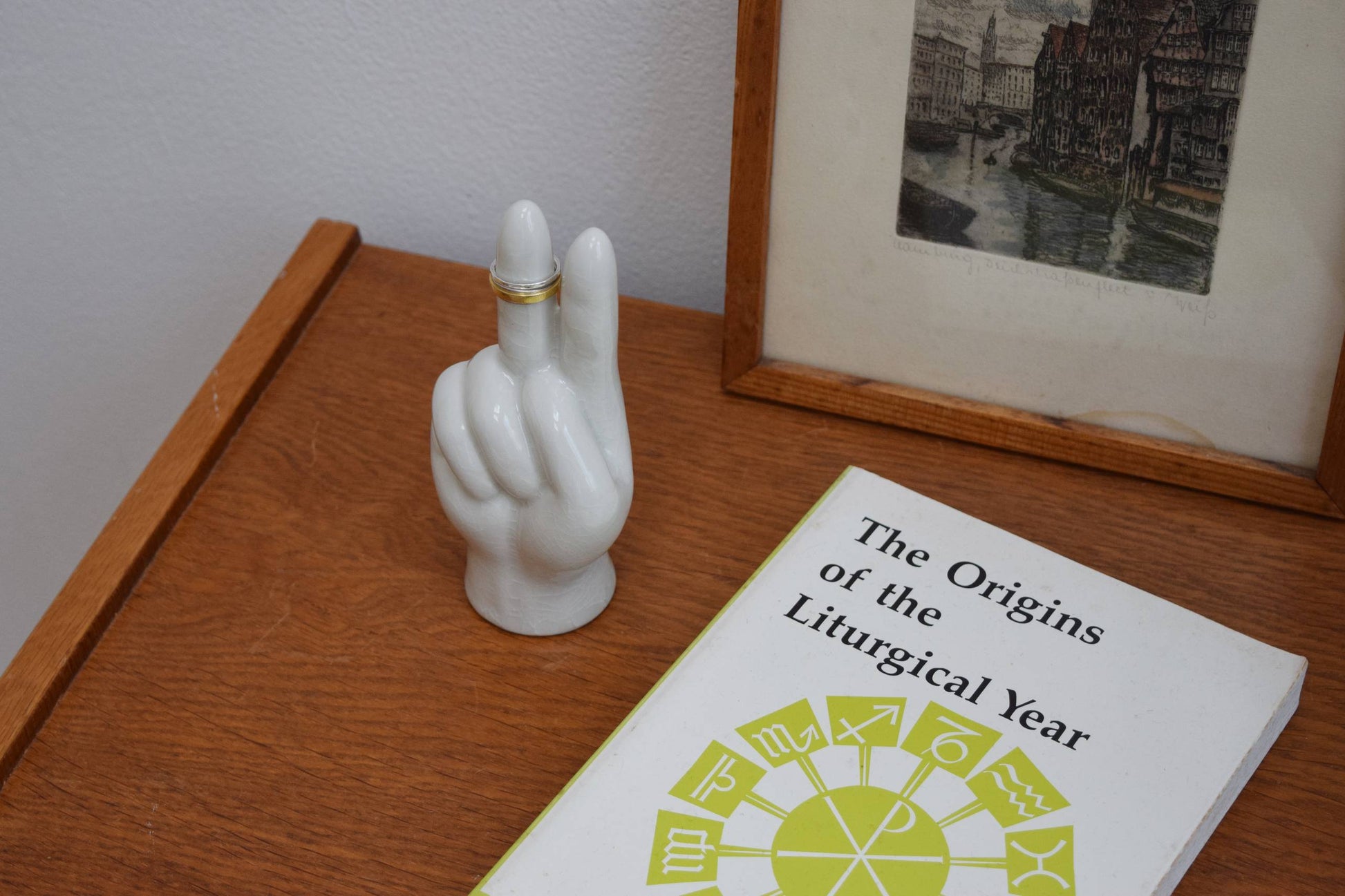 The DETAIL Porcelain Hand Peace by DETAIL elegantly holds a gold ring as an accessory stand, displayed on a wooden surface next to a framed photo and the book “The Origins of the Liturgical Year.”.