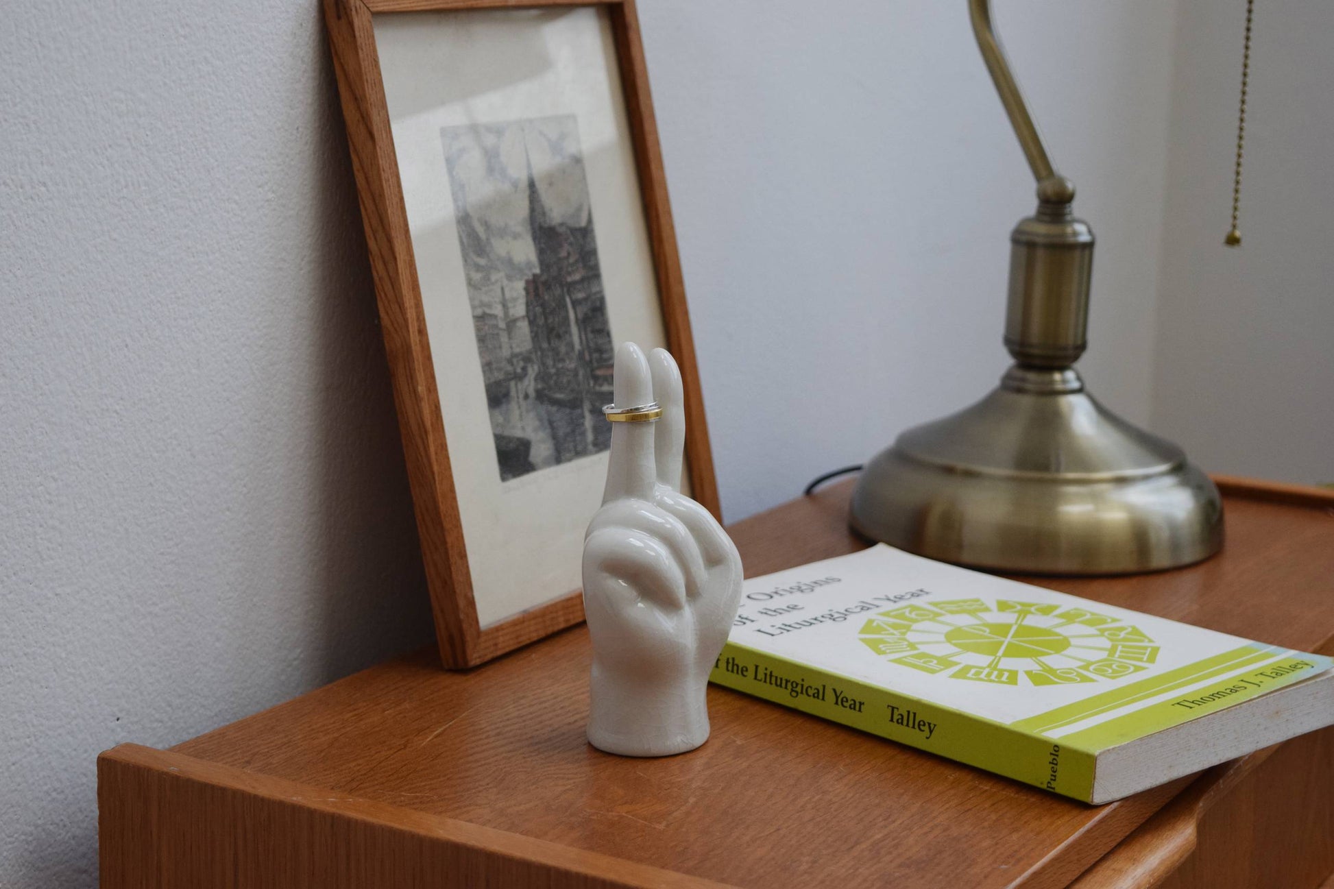 The DETAIL Porcelain Hand Peace by DETAIL holds two rings on a wooden nightstand next to a framed black-and-white photo, a brass lamp, and the book The Origins of the Liturgical Year.