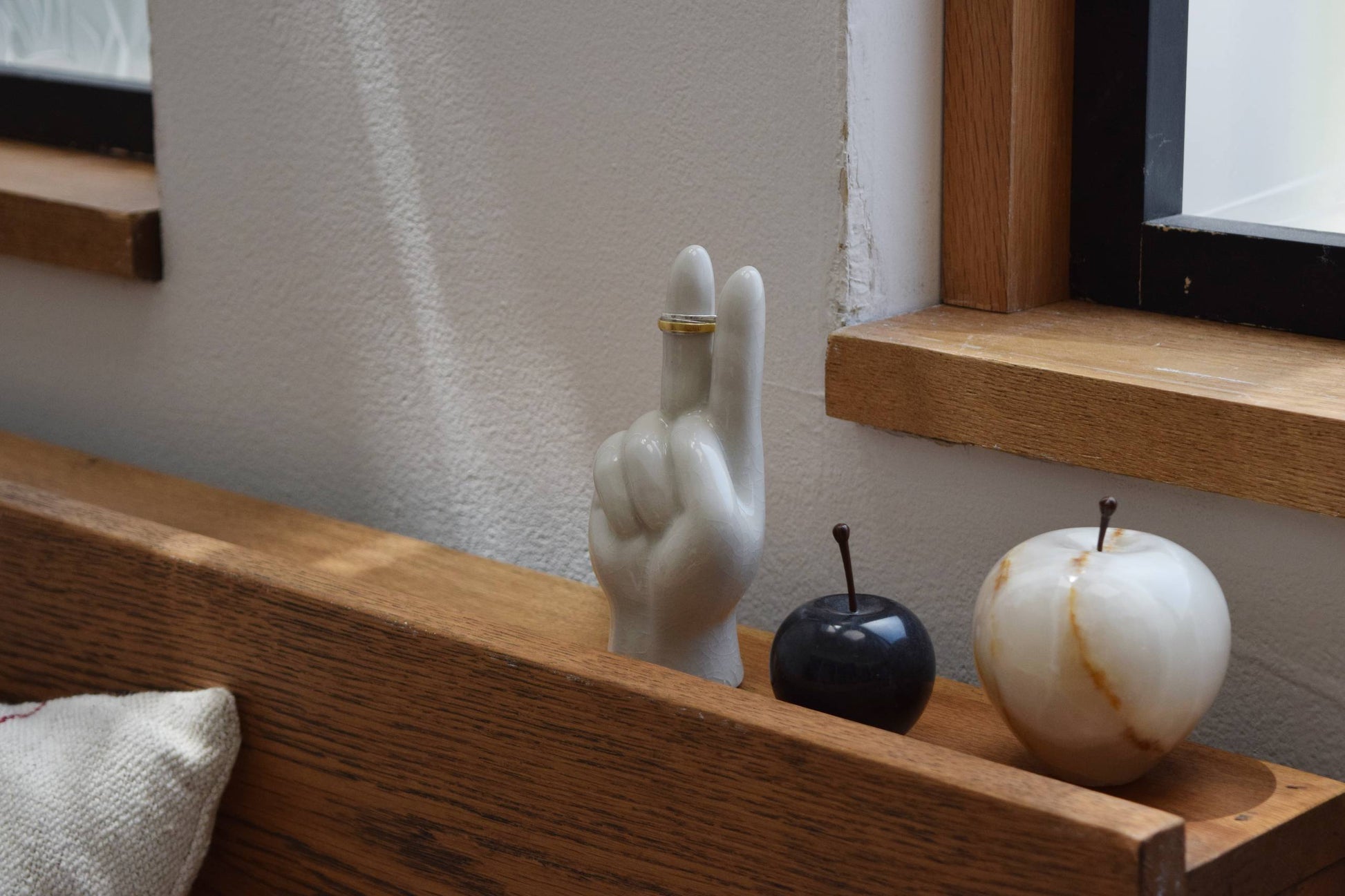The DETAIL Porcelain Hand Peace accessory stand, featuring a ring on its index finger, is displayed beside black and white apple ornaments on a wooden shelf by a window.