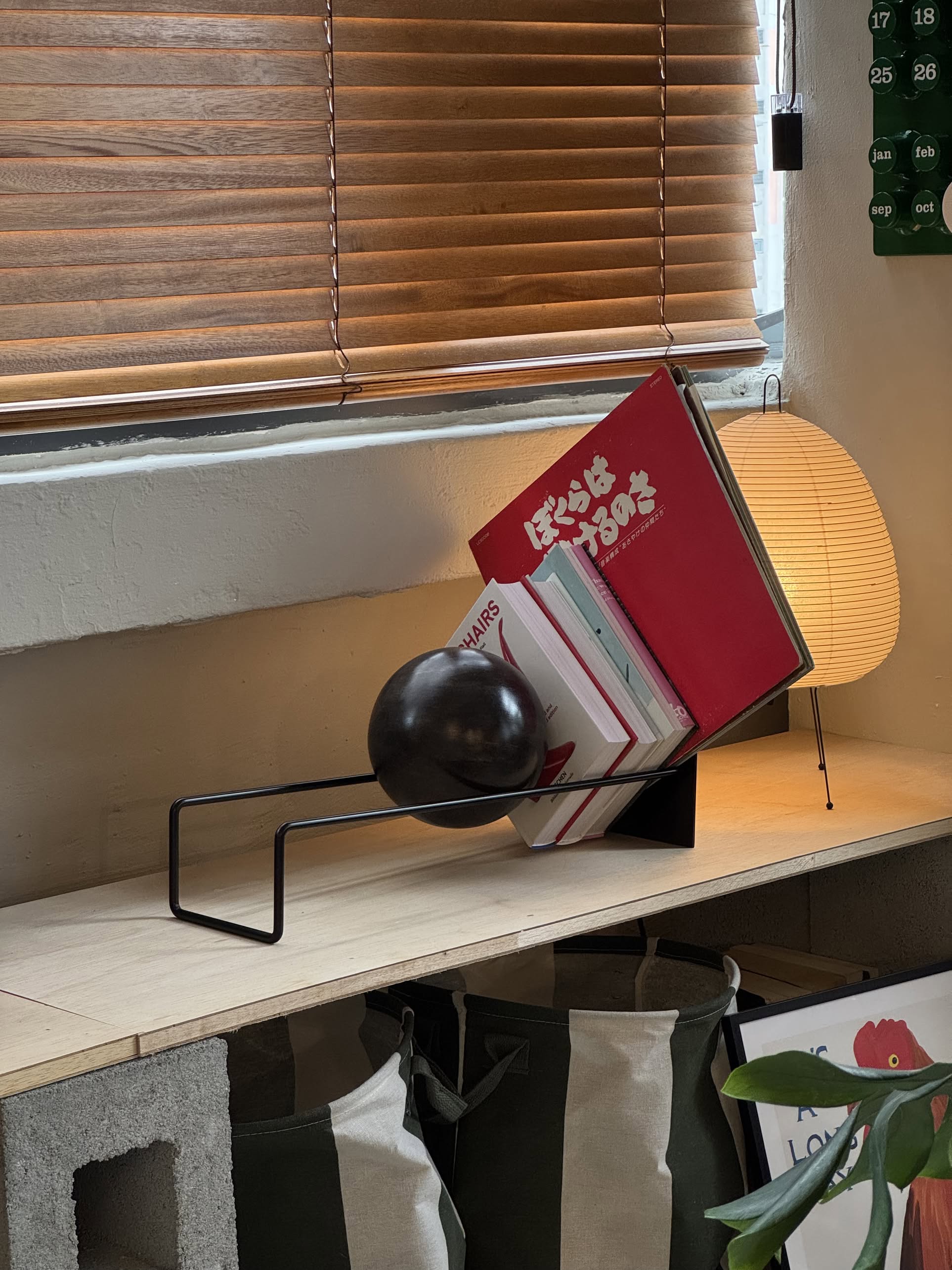 The DETAIL Slope Bookend by DETAIL, with its modern sculptural design, supports several books on a wooden shelf alongside a small lamp, blinds, and canvas storage bags.