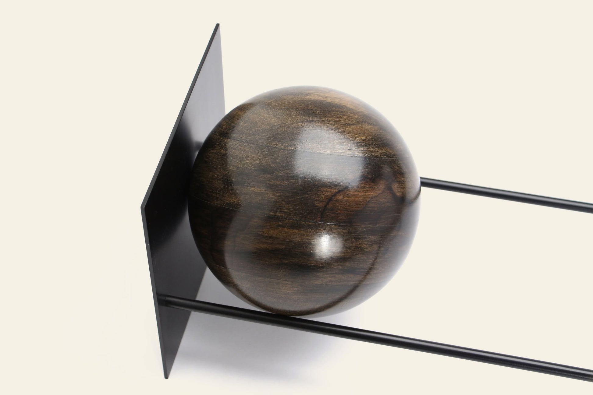 The DETAIL Slope Bookend by DETAIL features a polished wooden sphere against a vertical black metal plate, supported by two sleek parallel rods—a stylish minimalist accent on an off-white background.