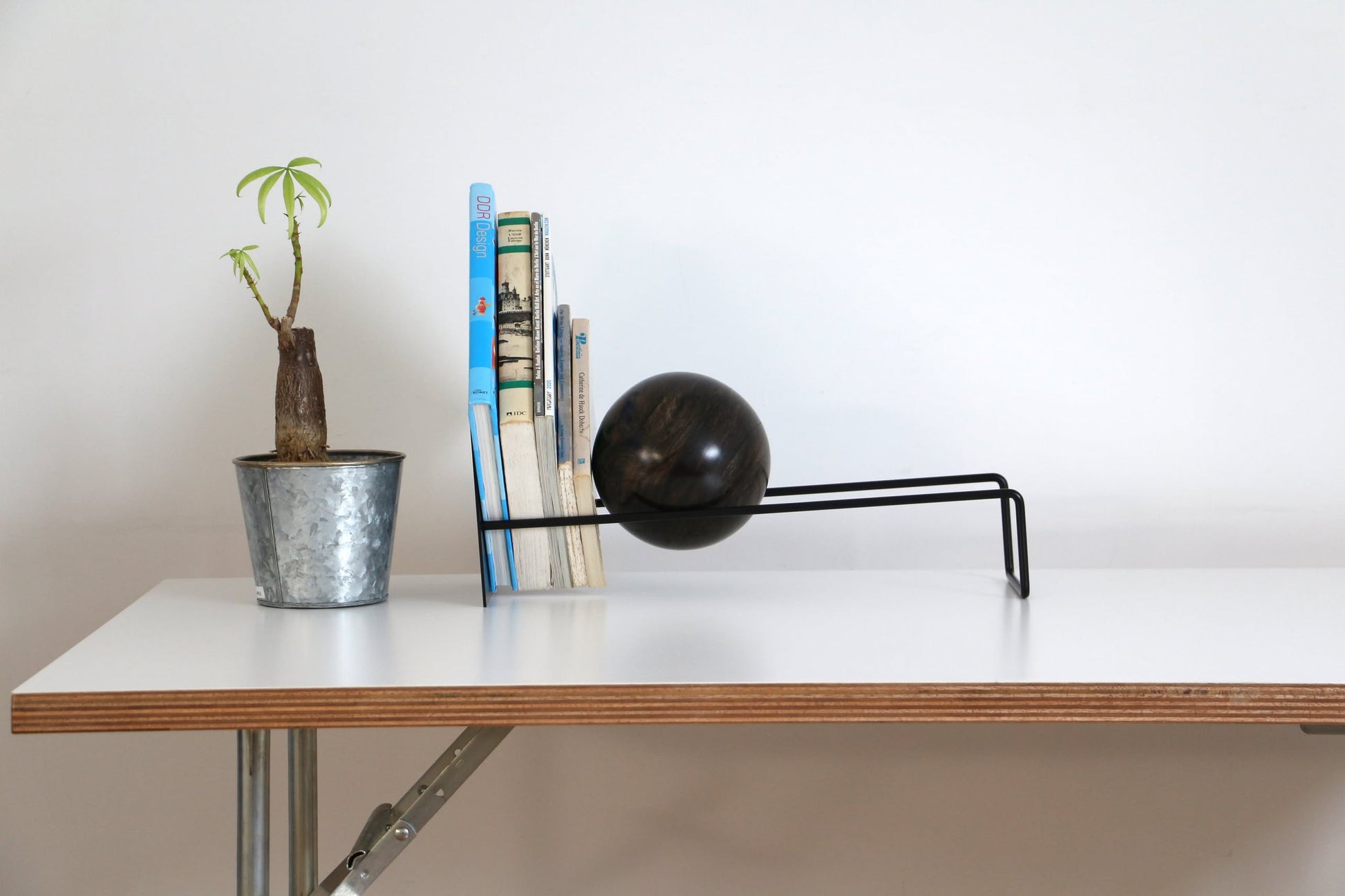 A small potted plant, several books, and the DETAIL Slope Bookend by DETAIL with a minimalist black metal design are arranged on a white desk against a plain white wall.