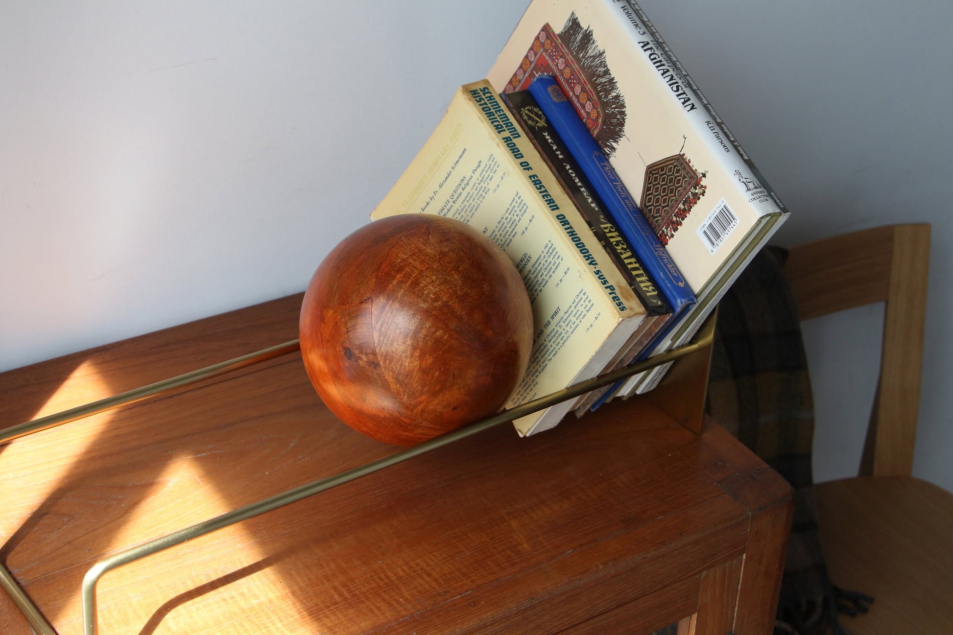 The DETAIL Slope Bookend by DETAIL features a unique mango wood ball holding books upright in a minimalist brass holder on wood, with sunlight casting shadows. A wooden chair and dark fabric appear in the background.