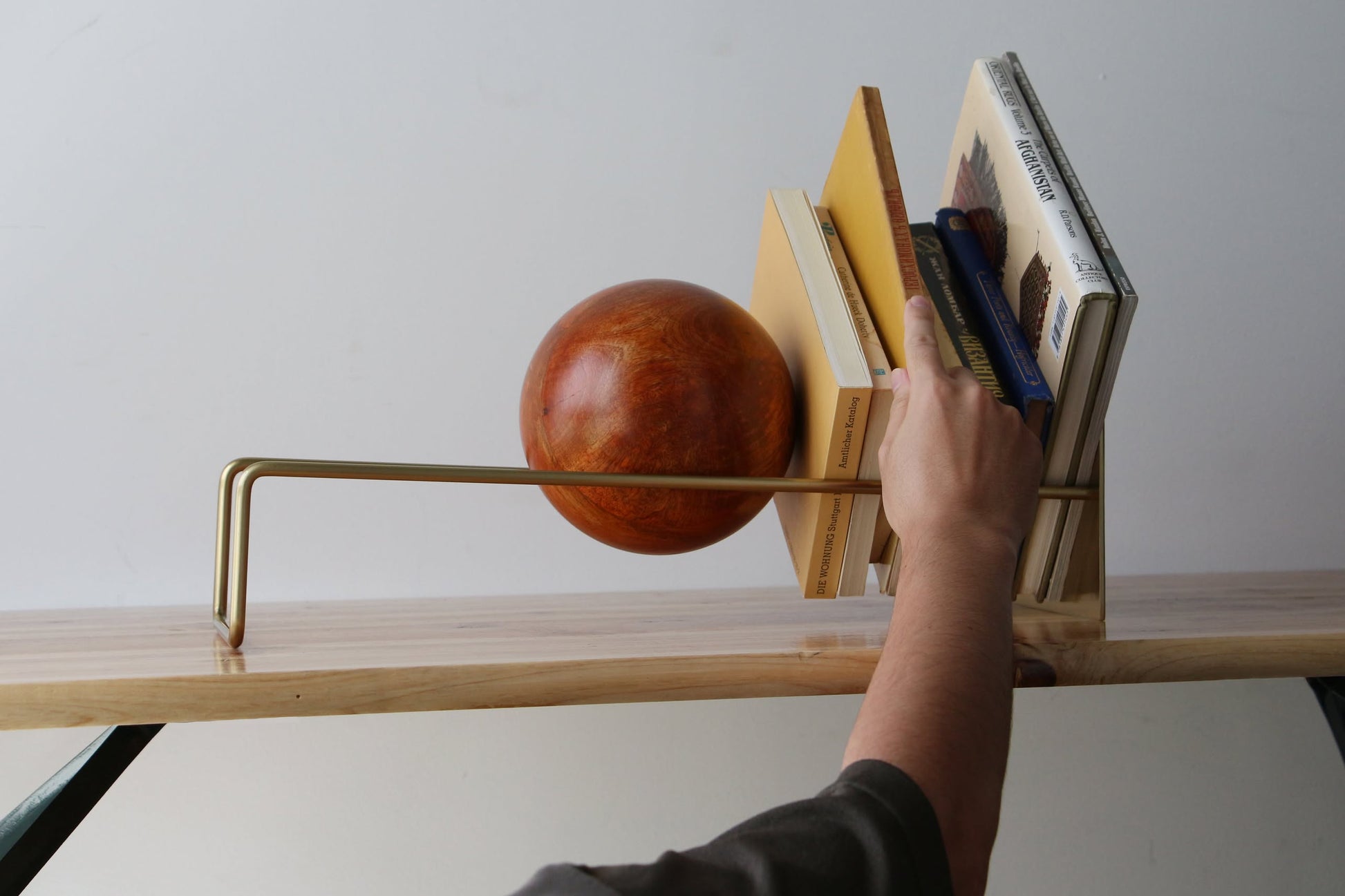 A hand arranges books on a modern bookshelf with minimalist design, featuring the DETAIL Slope Bookend by DETAIL in sleek silver steel and mango wood, set on light wood against a plain white background.