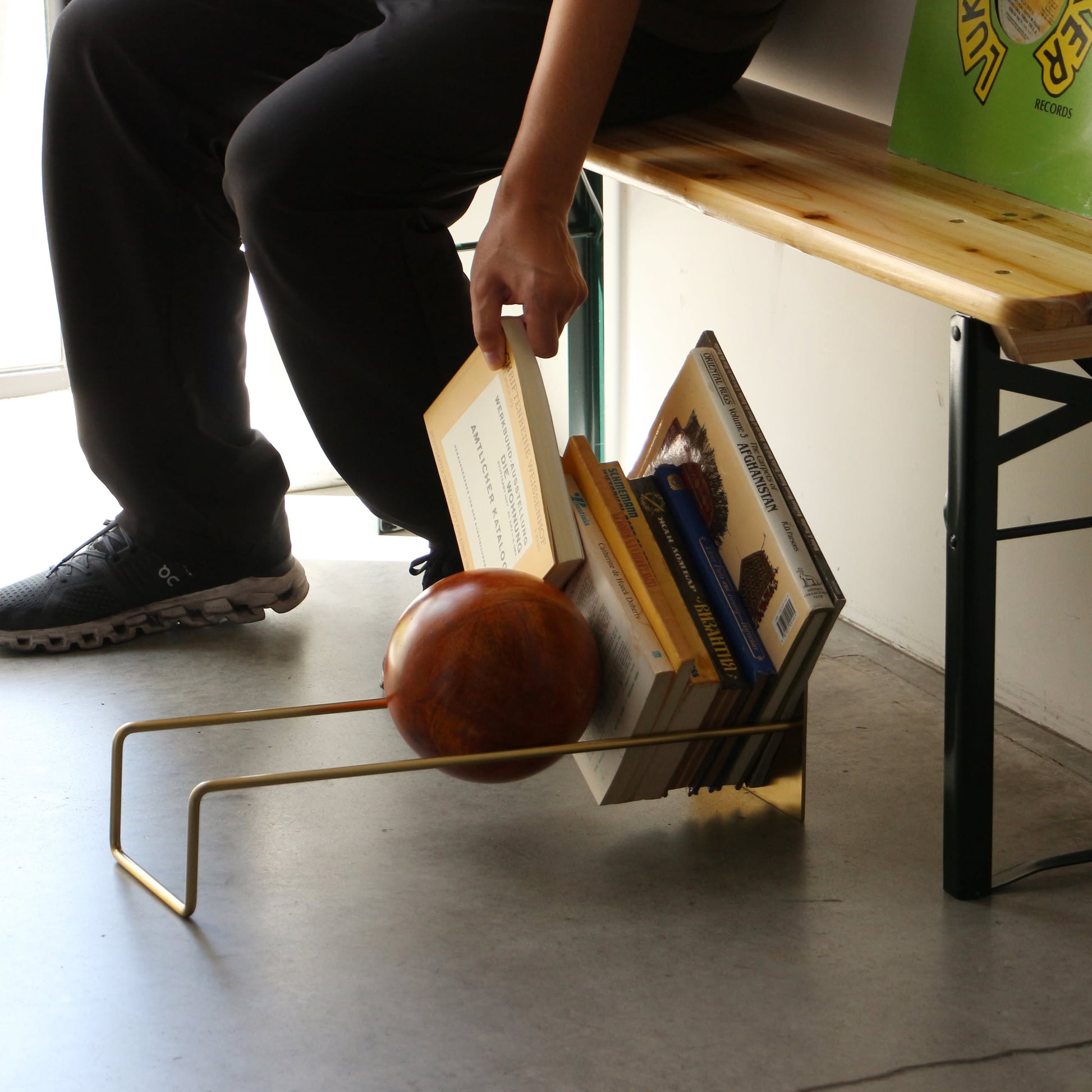 Sitting on a wooden bench, a person reaches for a book from the DETAIL Slope Bookend by DETAIL, which holds several books and a decorative mango wood ball on a concrete floor.