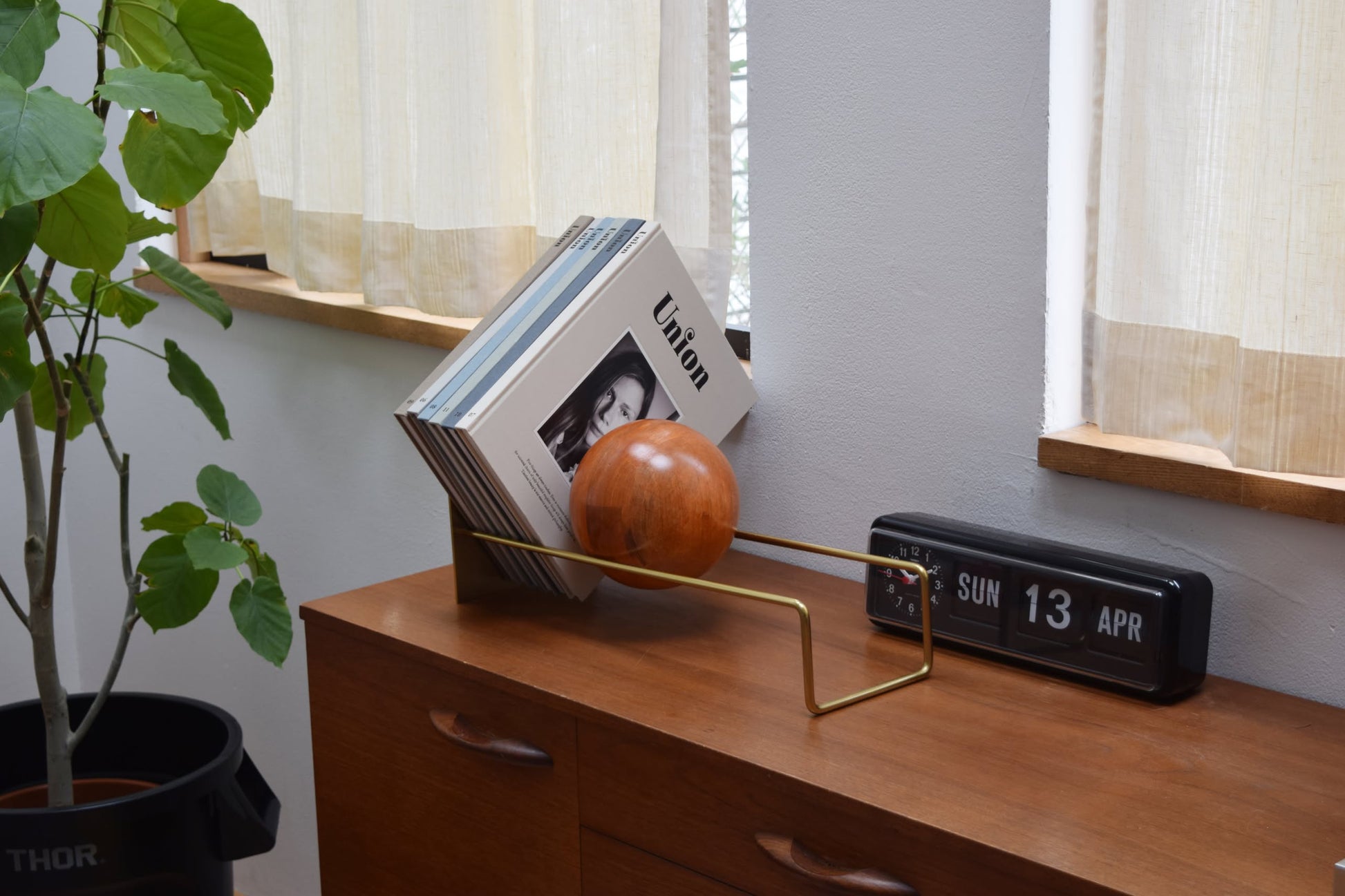 A stack of vinyl records is supported by the DETAIL Slope Bookend from DETAIL, featuring a unique mango wood ball. It’s displayed on a wooden sideboard near a black flip clock, with minimalist decor including a leafy plant and sheer curtains.