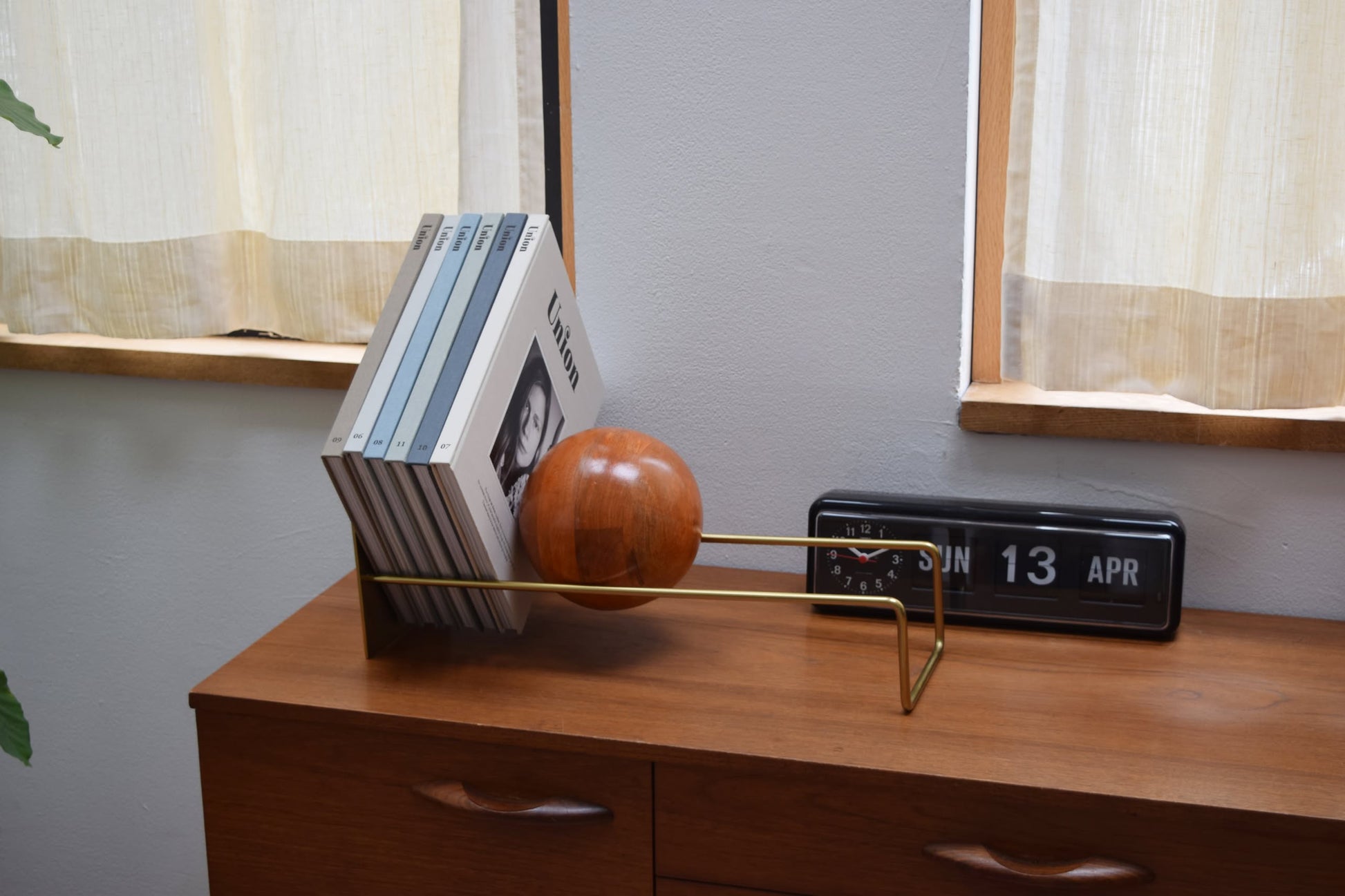 A modern wooden sideboard displays the DETAIL Slope Bookend by DETAIL holding magazines and a black flip clock set to “SUN 13 APR,” perfectly placed between two windows with light curtains.