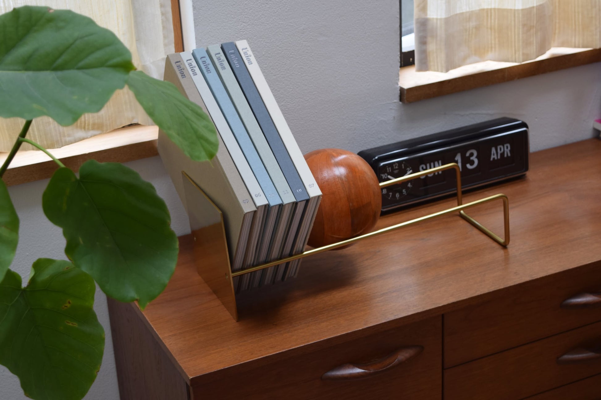 A wooden dresser displays the DETAIL Slope Bookend by DETAIL with books, a mango wood ball, a vintage flip clock showing SUN 13 APR, and a leafy plant near a sunlit window.