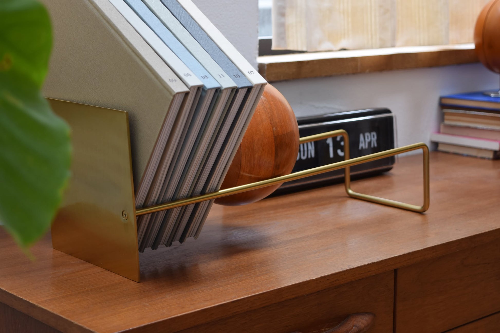 The DETAIL Slope Bookend by DETAIL features a modern brass design with a sleek silver steel accent, keeping books upright on a wooden desk. A retro flip clock displays SUN 13 APR, and small books sit by a sunlit window nearby.