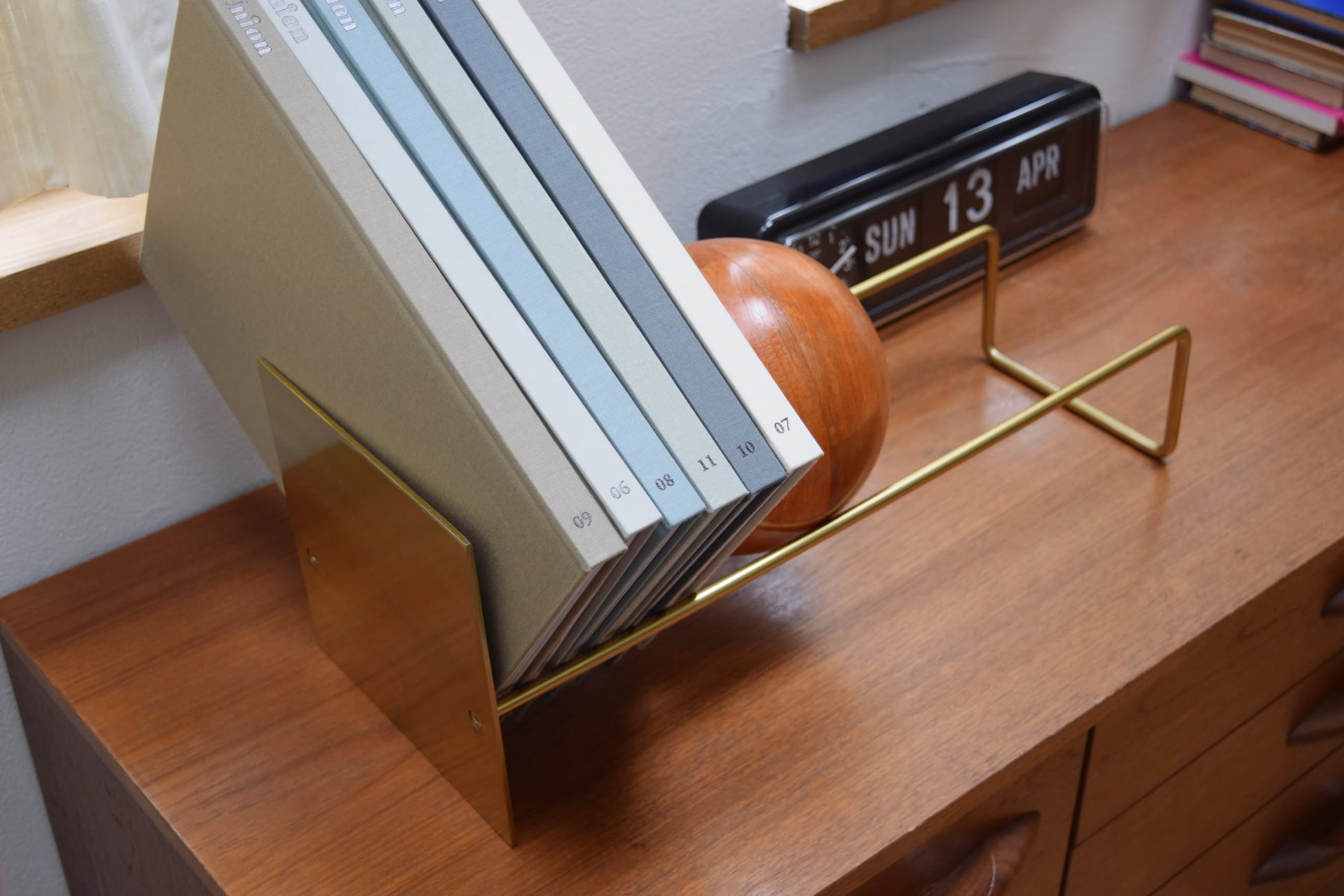 The DETAIL Slope Bookend by DETAIL holds several neatly arranged books with numbered spines on a wooden desk, accompanied by a distinctive mango wood ball and a black retro flip clock showing Sunday, April 13.