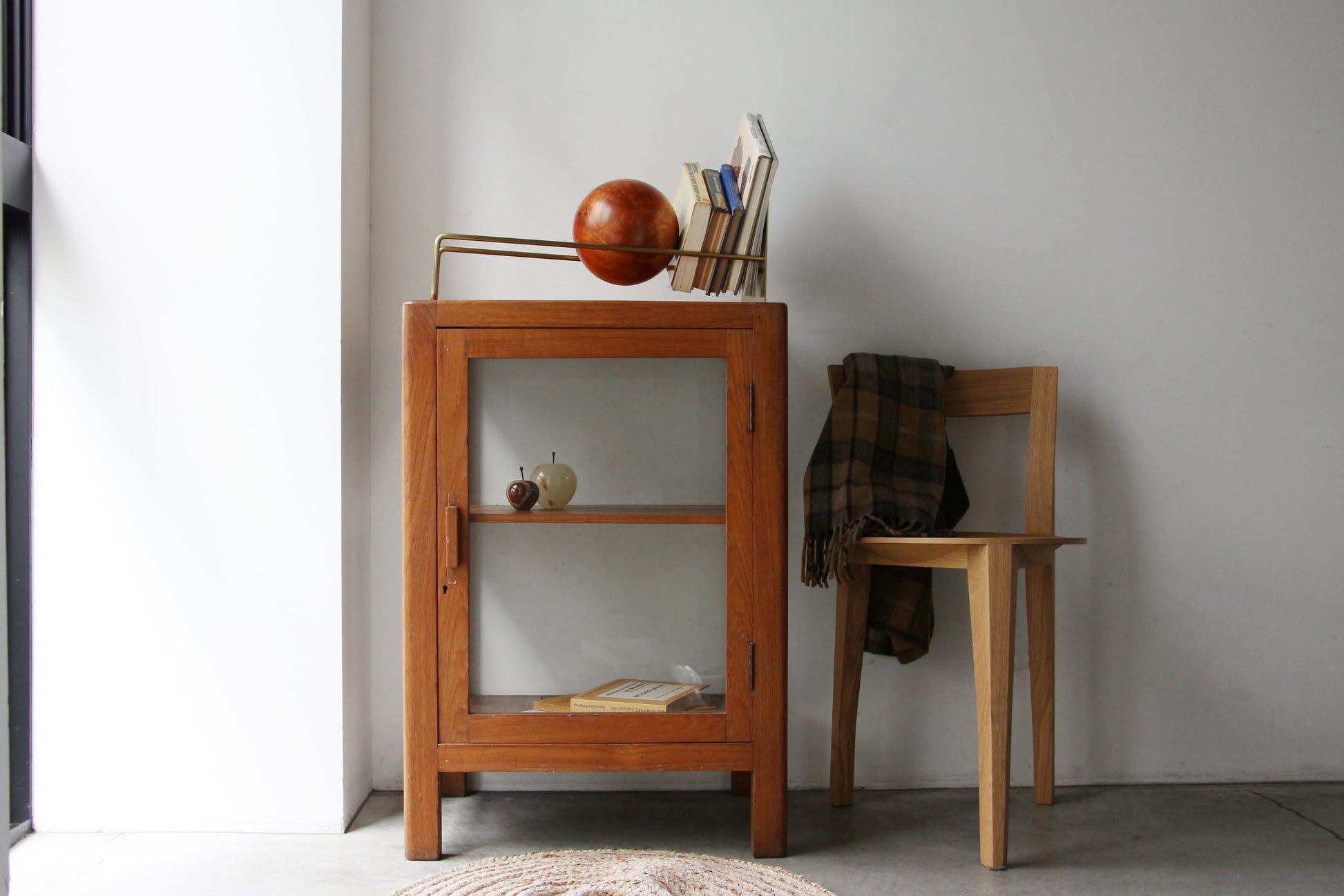 A DETAIL Slope Bookend by DETAIL sits atop a small wooden cabinet with a glass door beside a simple chair draped in a plaid blanket, enhancing the minimalist look of this bright, understated space.