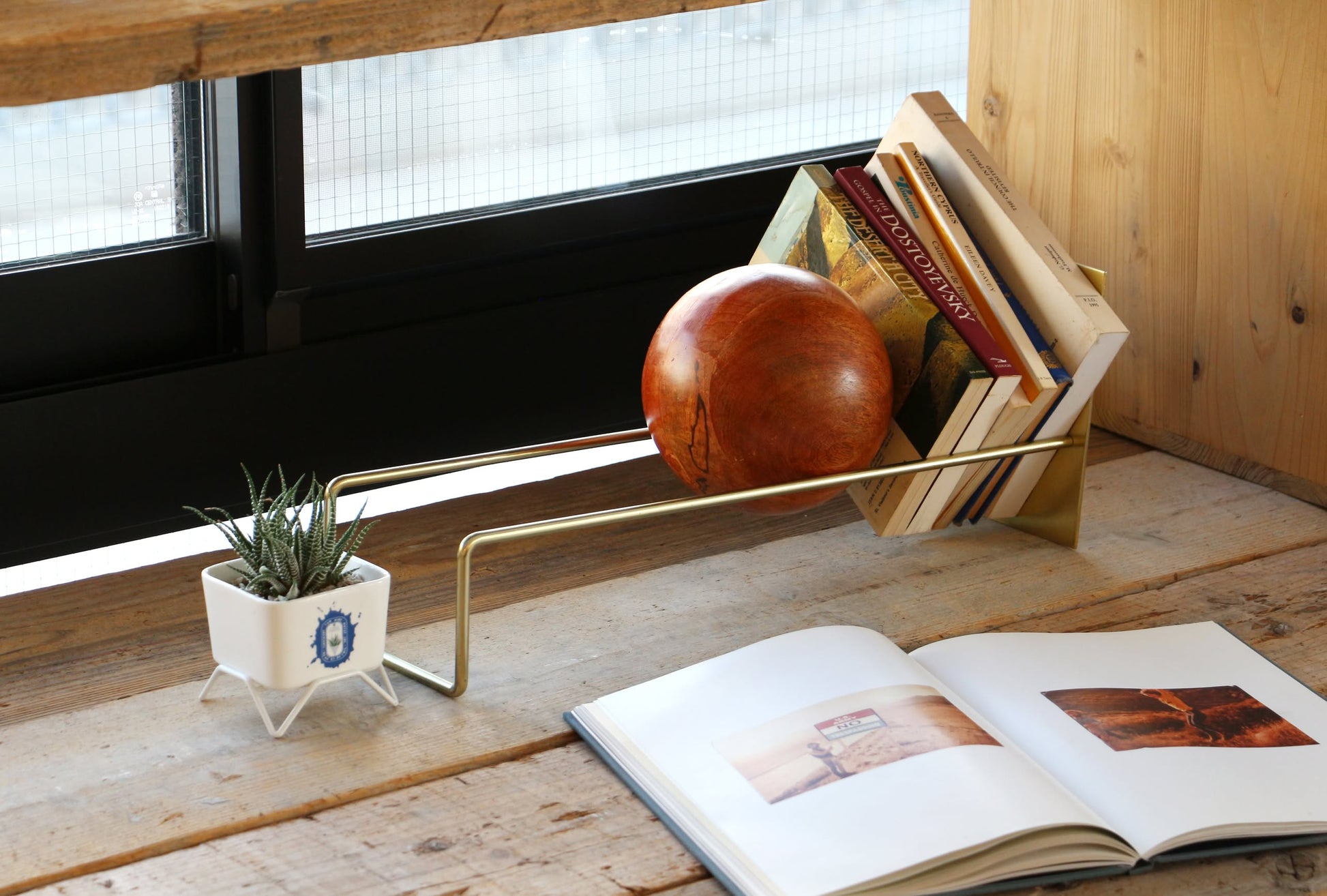A DETAIL Slope Bookend by DETAIL organizes several books and a unique mango wood ball on a modern gold shelf. A small potted succulent sits at one end, while an open photo book lies on a wooden table in front of black-framed windows.