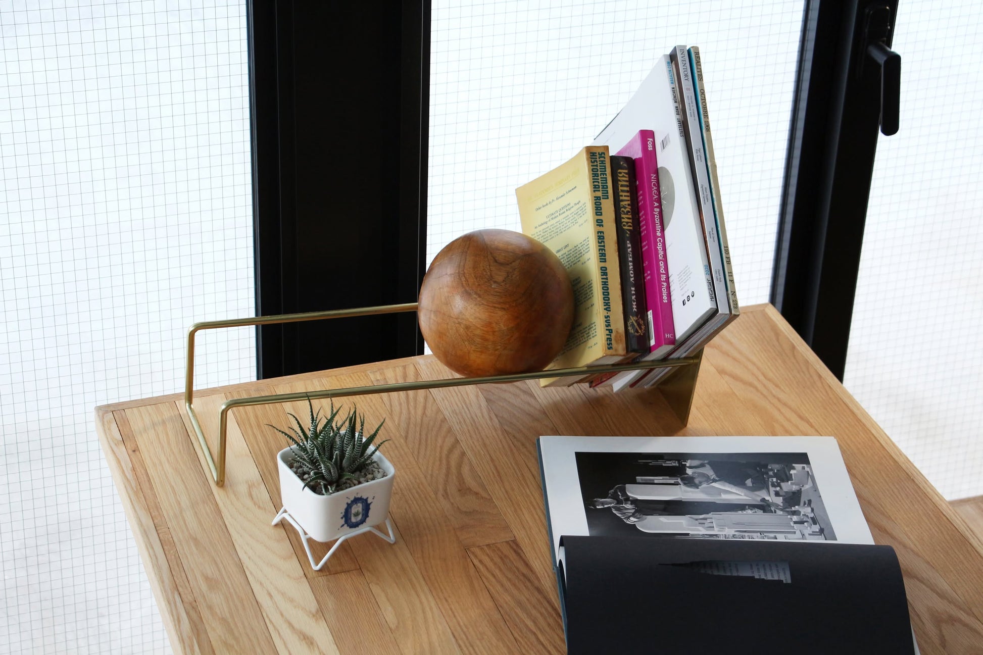 The DETAIL Slope Bookend by DETAIL holds books and a distinctive mango wood ball on a tabletop, next to a small potted succulent and an open book with black-and-white photos, all set near large windows.