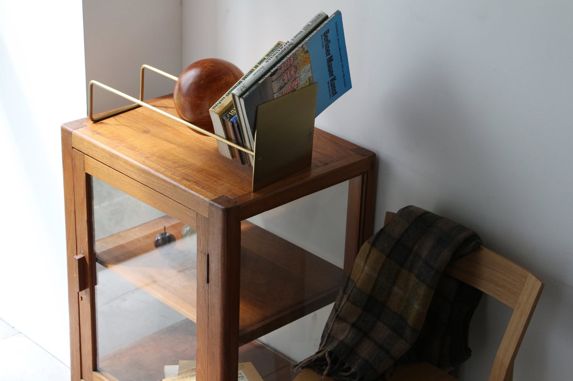 A wooden cabinet with glass doors stands beside a wooden chair draped with a plaid scarf. On the cabinet, the DETAIL Slope Bookend by DETAIL holds books and magazines, while a unique mango wood ball adds to the minimalist design.