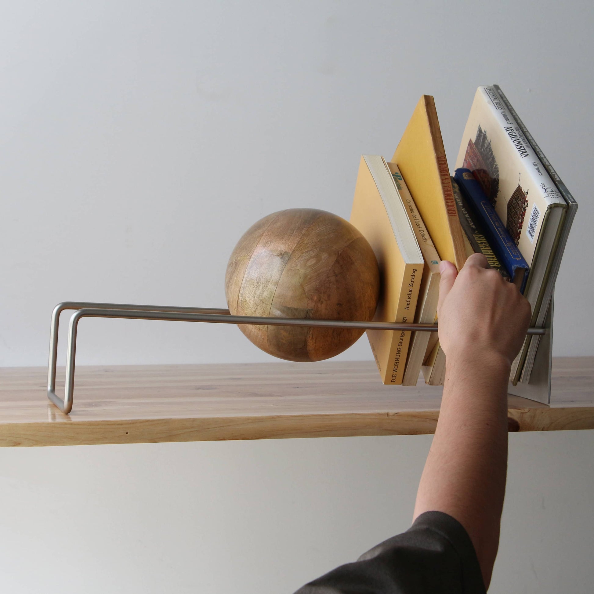 A hand reaches for a book on the DETAIL Slope Bookend by DETAIL, displayed with several books and a distinctive mango wood ball, all arranged on a wooden surface against a plain, light background.