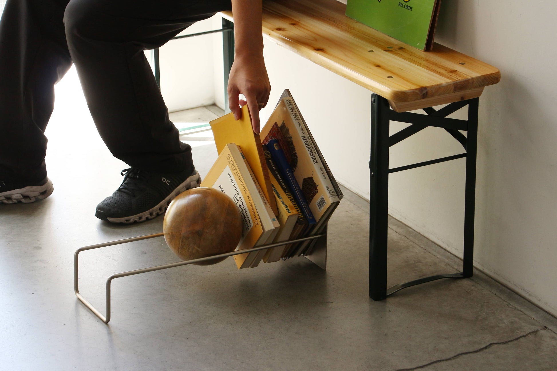 A person in black pants and sneakers picks up a yellow folder from the DETAIL Slope Bookend by DETAIL, a sleek silver steel book rack on the floor holding books and a unique mango wood ball.