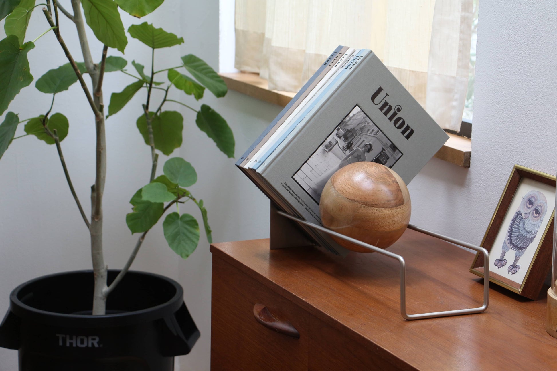 A wooden sideboard displays the DETAIL Slope Bookend by DETAIL, holding magazines and a mango wood ball, beside a framed owl illustration. A potted plant by a bright window accentuates the minimalist decor.