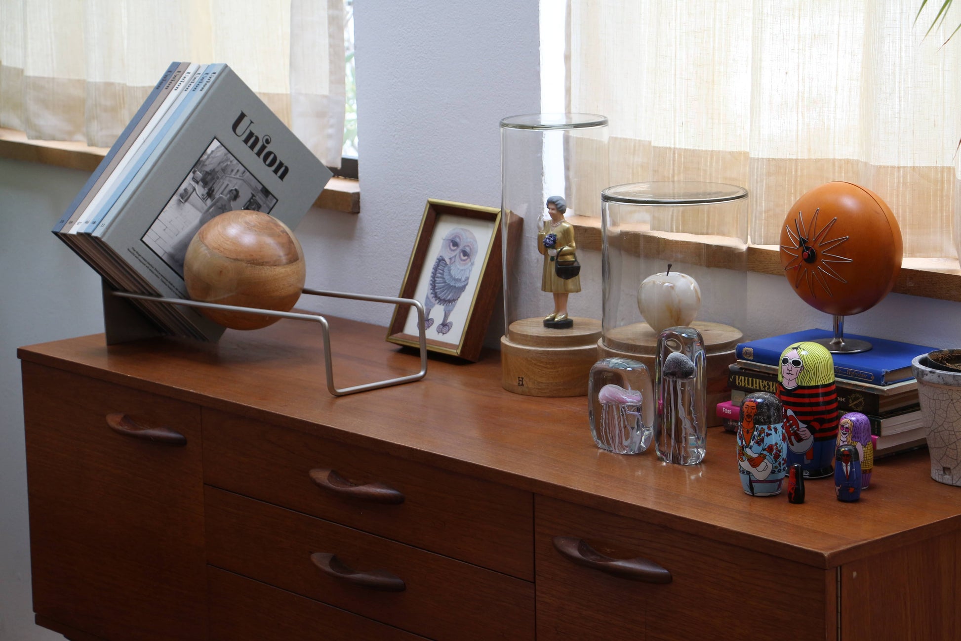 A wooden dresser with minimalist design displays books held by DETAIL Slope Bookend from DETAIL, a mango wood ball sculpture, framed owl illustration, glass domes with figures inside, nesting dolls, and more. Sunlight filters through sheer curtains.