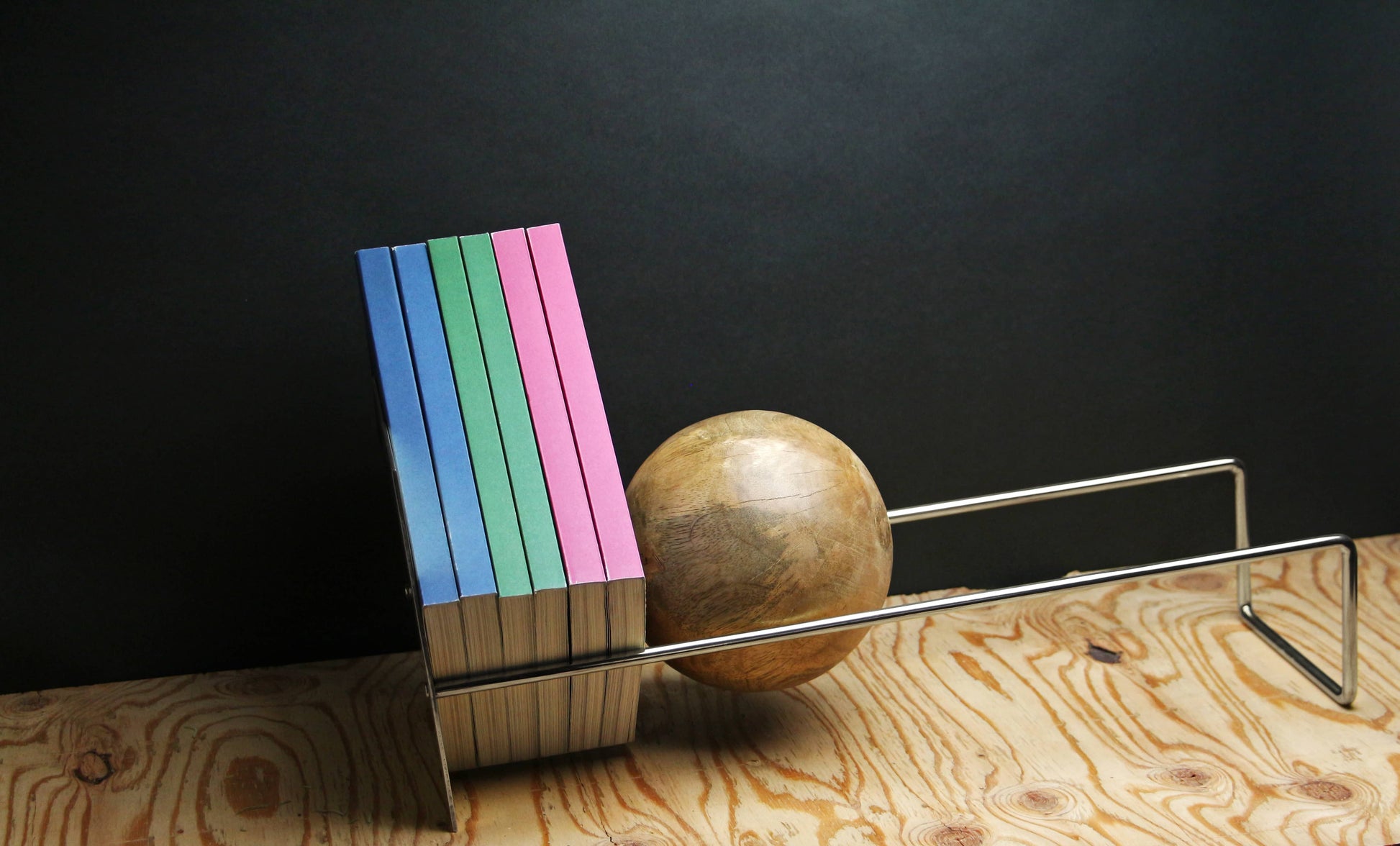Six pastel-colored books stand upright on a wooden surface, supported by the DETAIL Slope Bookend from DETAIL. Its sleek silver steel design features a unique mango wood ball, creating a modern, minimalist look against the dark background.