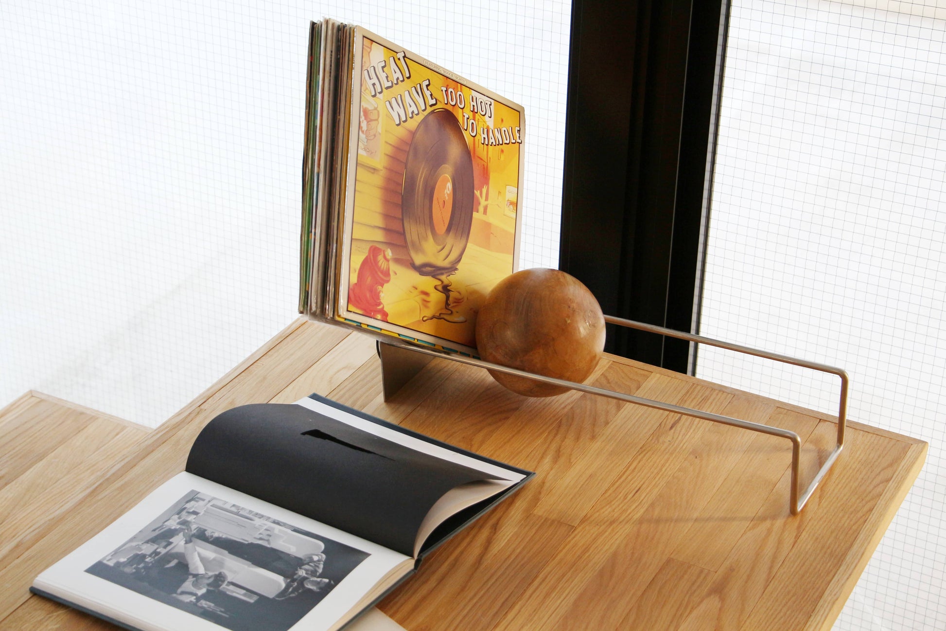 The DETAIL Slope Bookend by DETAIL, featuring a sleek silver steel frame and a mango wood ball, displays vinyls. Nearby, an open black-and-white photo book sits on a wooden table next to a grid-patterned window.