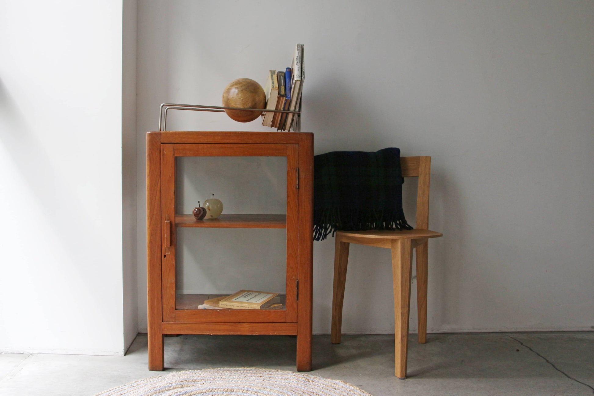 A small wooden cabinet with a glass door sits beside a wooden chair draped in a dark plaid blanket. Atop the cabinet, the DETAIL Slope Bookend by DETAIL and books add charm to the minimalist design against a plain white wall.